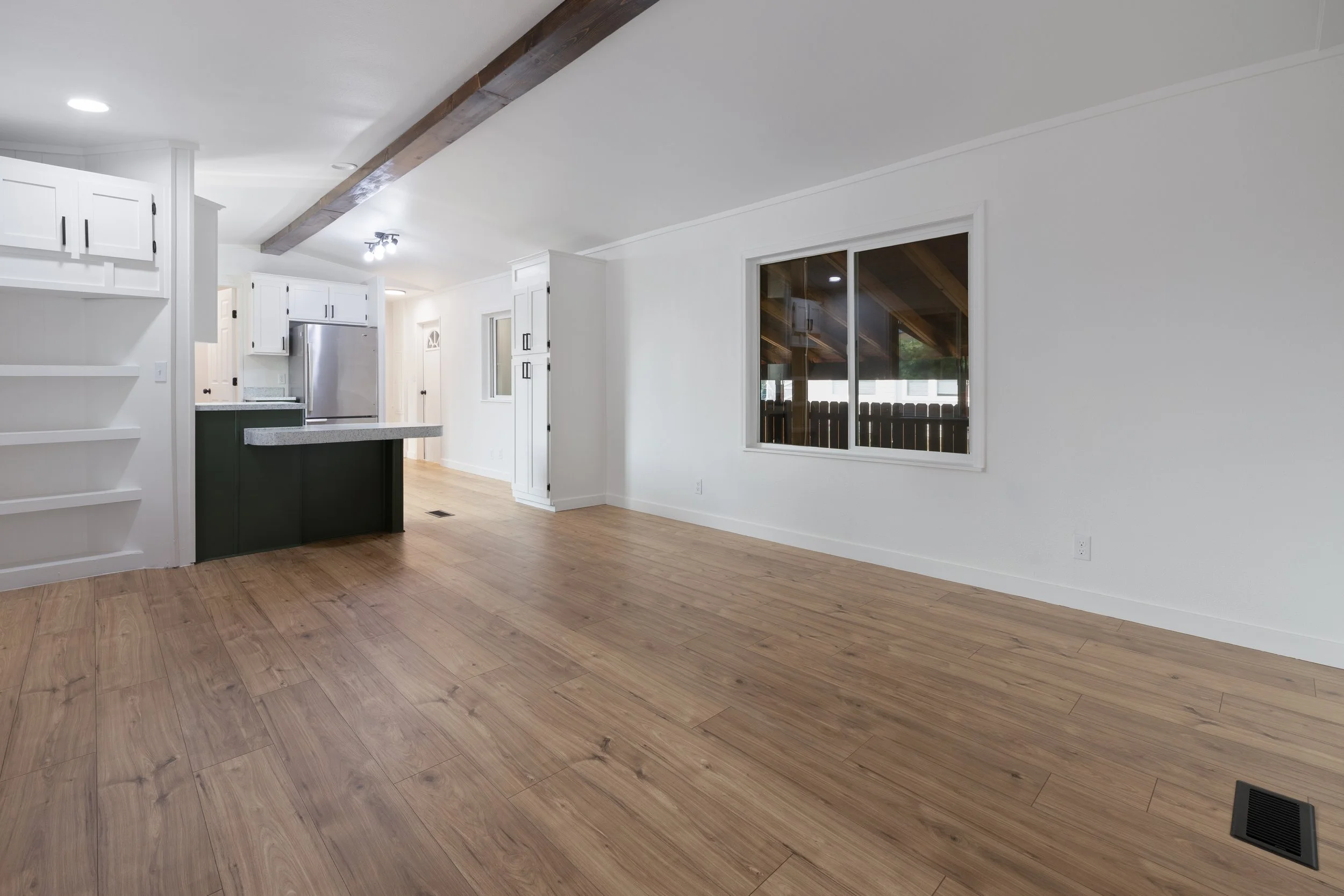 Empty living room with light wood flooring, white walls, a large window, and a partial view of a kitchen with white cabinets and a gray countertop.