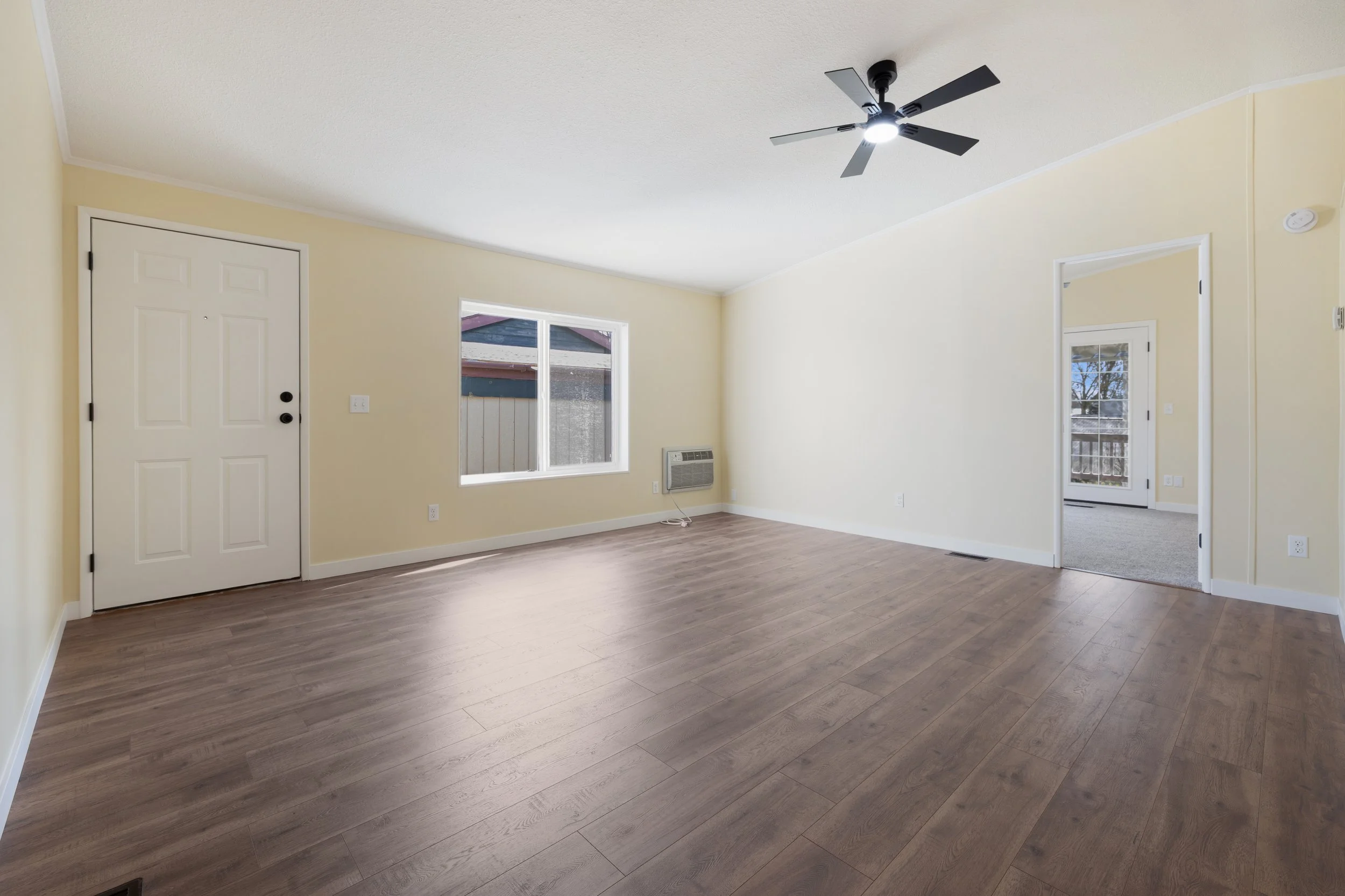 Empty living room with beige walls, a ceiling fan, two windows, and a door leading to a deck. Wood flooring covers the space.