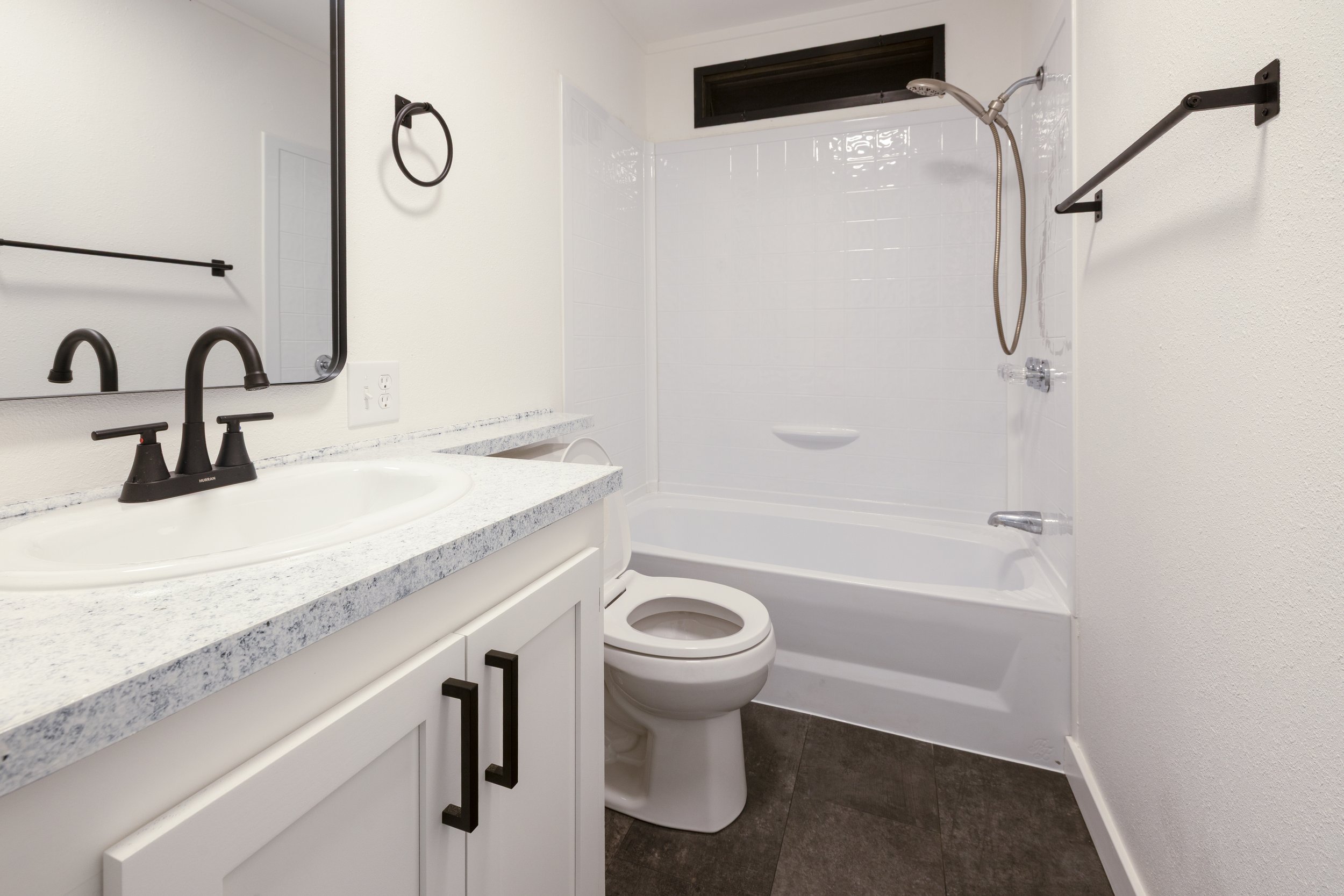 A small bathroom with a white vanity, a black faucet, a mirror, a toilet, a bathtub with a showerhead, a small window, a towel ring, and a towel rod.
