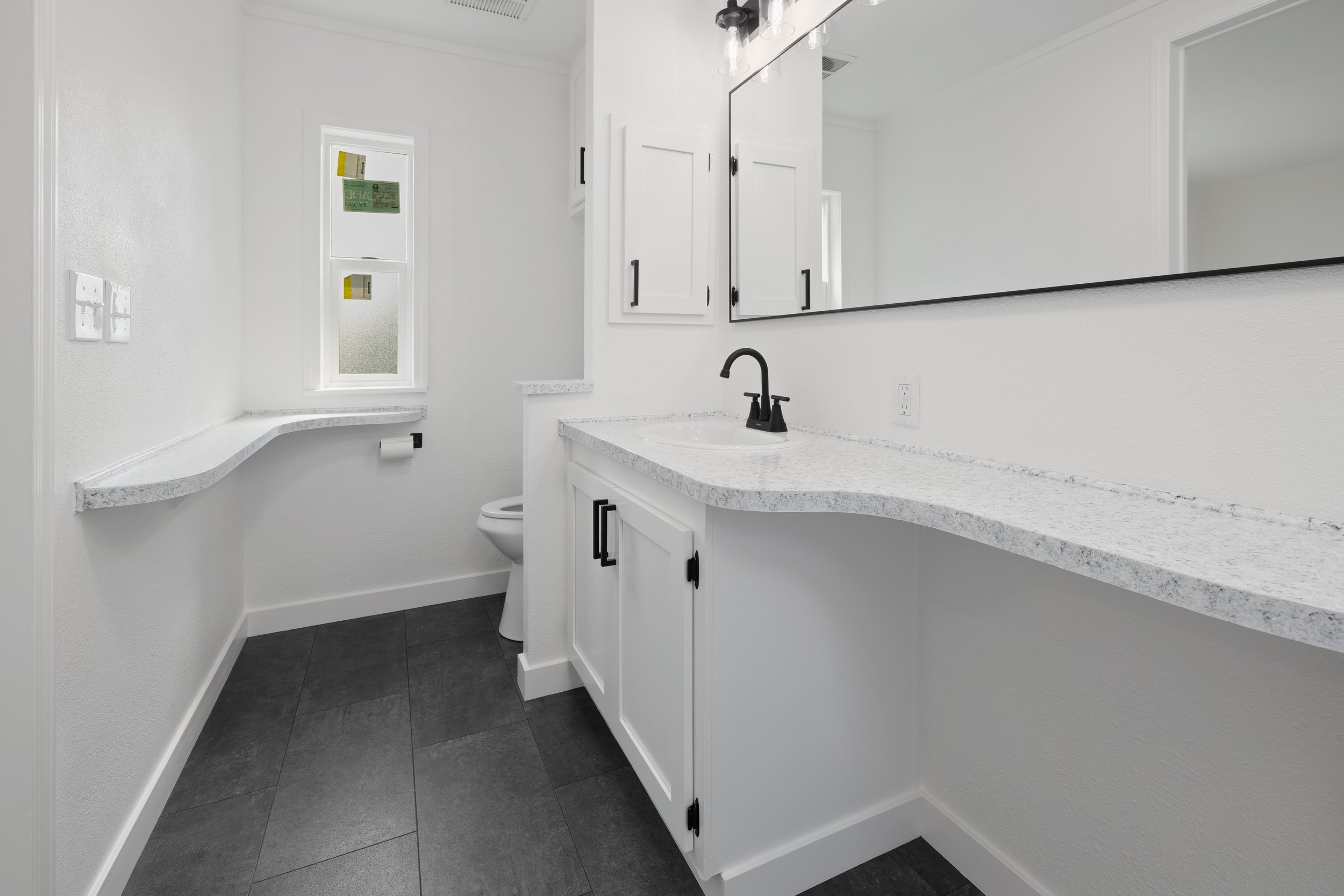 Modern white bathroom with black fixtures, a large mirror, gray tile floor, and a vanity with a speckled countertop.