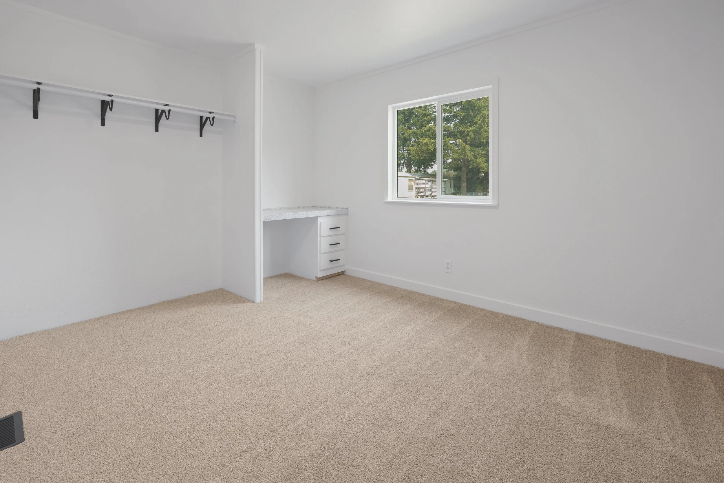 Empty room with beige carpet, white walls, a window showing trees outside, a built-in desk, and a coat rack shelf.