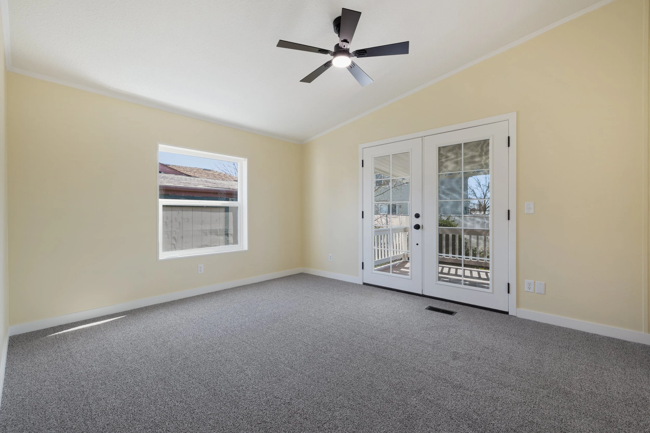 Empty living room with yellow walls, cream-colored carpet, double glass-paneled door leading to a balcony, and a ceiling fan with lights on the ceiling.
