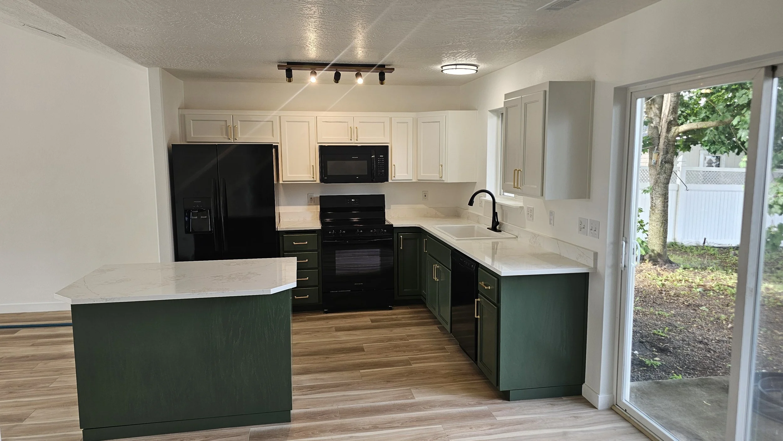 Modern kitchen with black appliances, white upper cabinets, dark green lower cabinets with gold handles, white marble countertops, a sink near a window, and a sliding glass door leading outside to a yard with a tree and fencing.