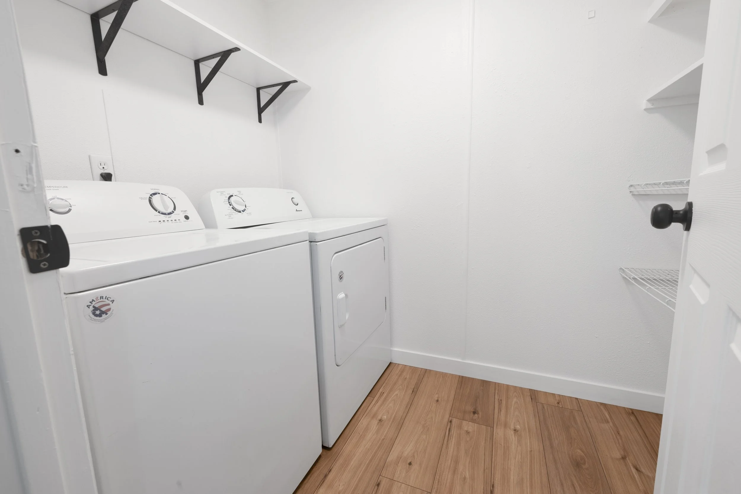 A small laundry room with white walls and wood laminate flooring, featuring a white washing machine and dryer, black wall shelves, and an open door with a wire storage shelf attached.