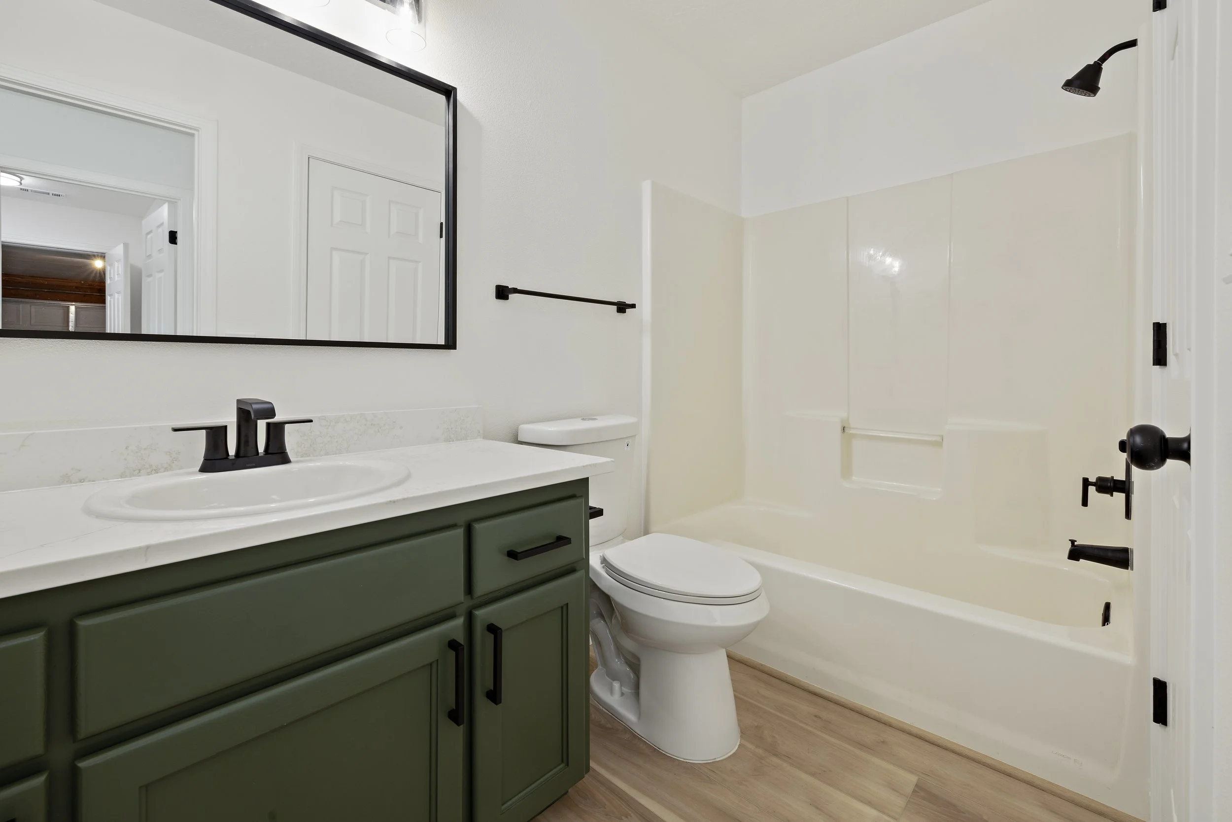 A bathroom featuring a green vanity cabinet with black handles, a white countertop with a built-in sink and black faucet, a toilet, a large mirror with a black frame, and a shower with a black showerhead and fixtures, all with light-colored walls and wood-look flooring.