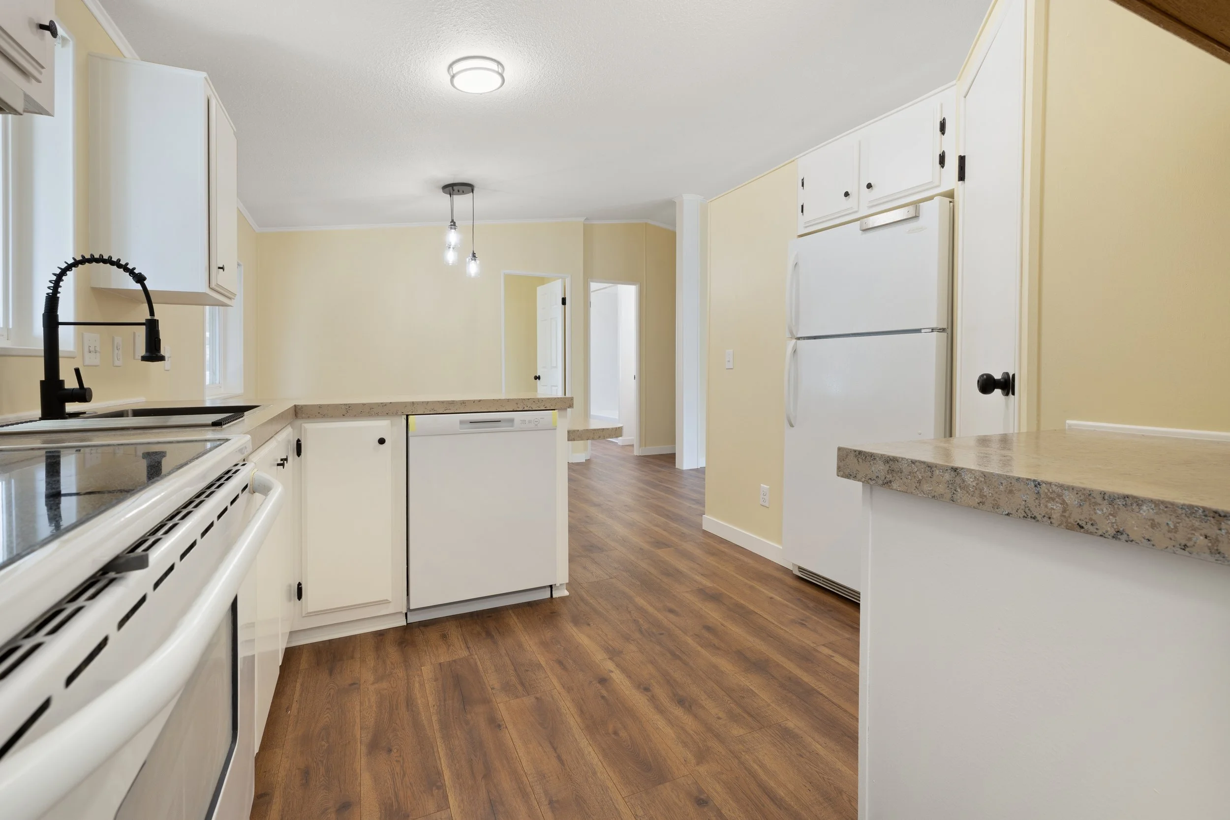 A kitchen with white cabinets, a beige granite countertop, a white refrigerator, a black sink faucet, electric stove, and wooden flooring. The kitchen has two doorways and an overhead ceiling light fixture.