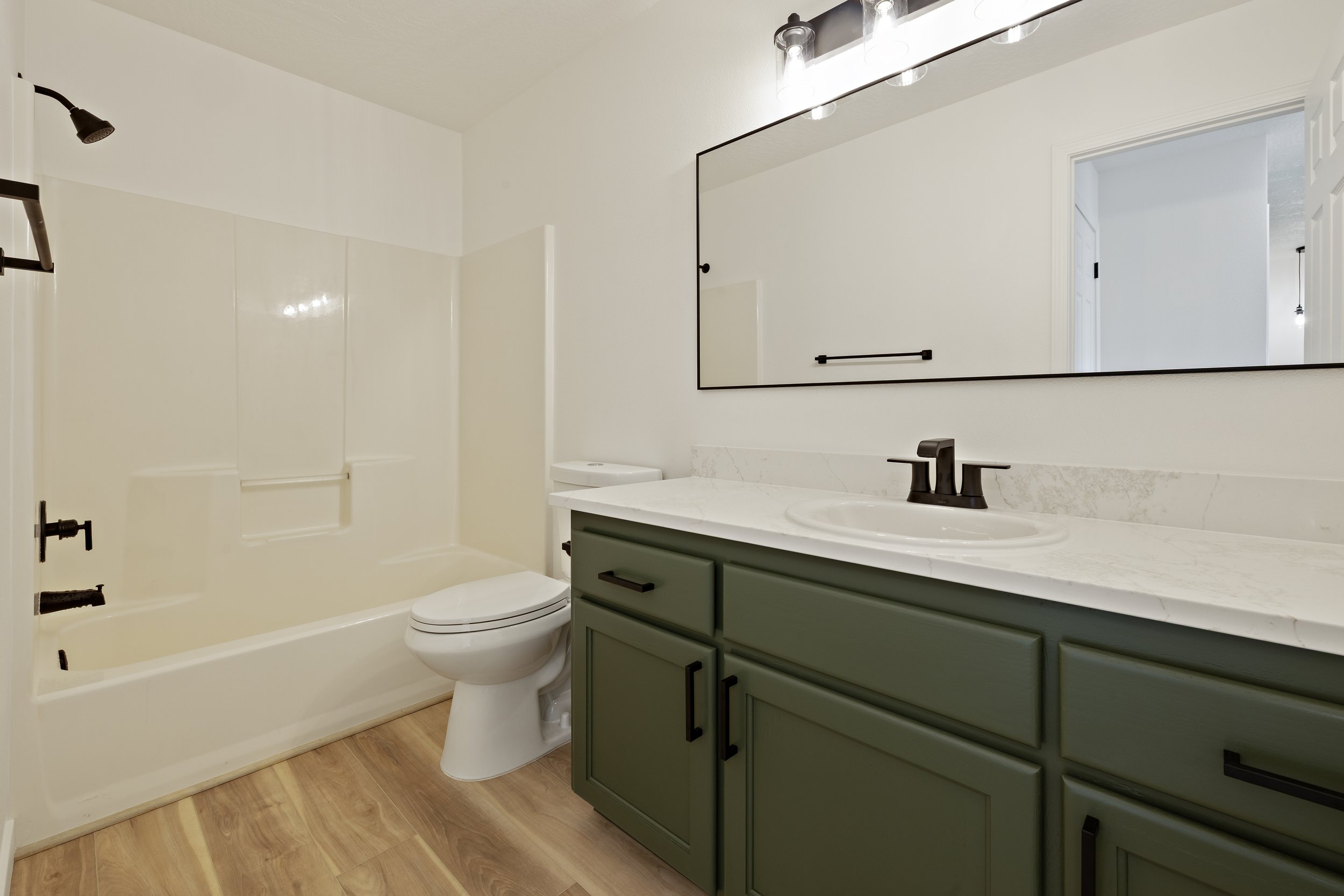 Modern bathroom with a white bathtub and toilet, a green vanity with black hardware, a marble countertop, and a large mirror above the sink. The bathroom has light wood flooring and a black light fixture.