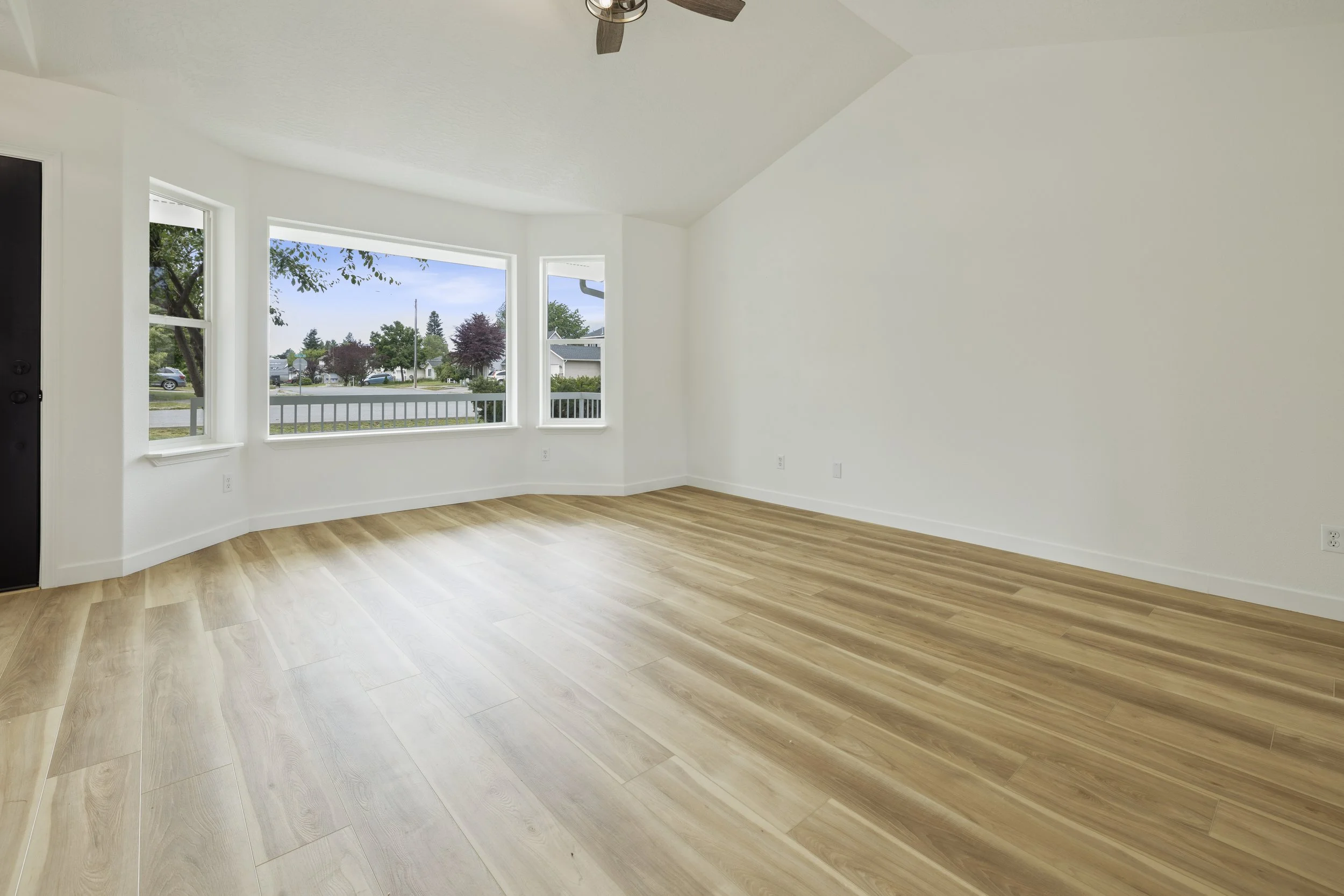 Empty room with light wood flooring, white walls, three large windows, and a ceiling fan.