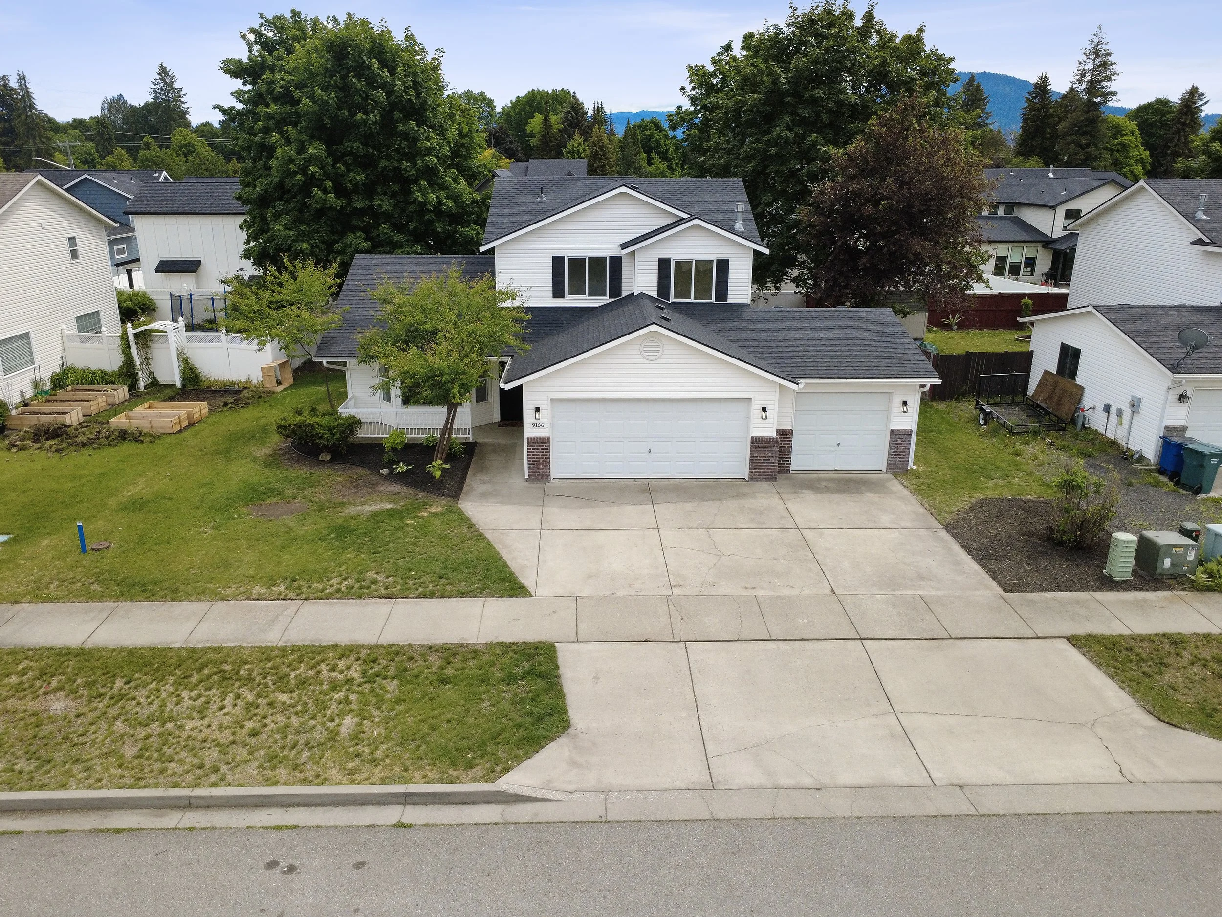 A two-story white house with a three-car garage, a well-maintained lawn, and surrounding trees. There is a sidewalk in front of the house, and neighboring houses are visible on either side. In the background, there are green hills and trees under a clear blue sky.