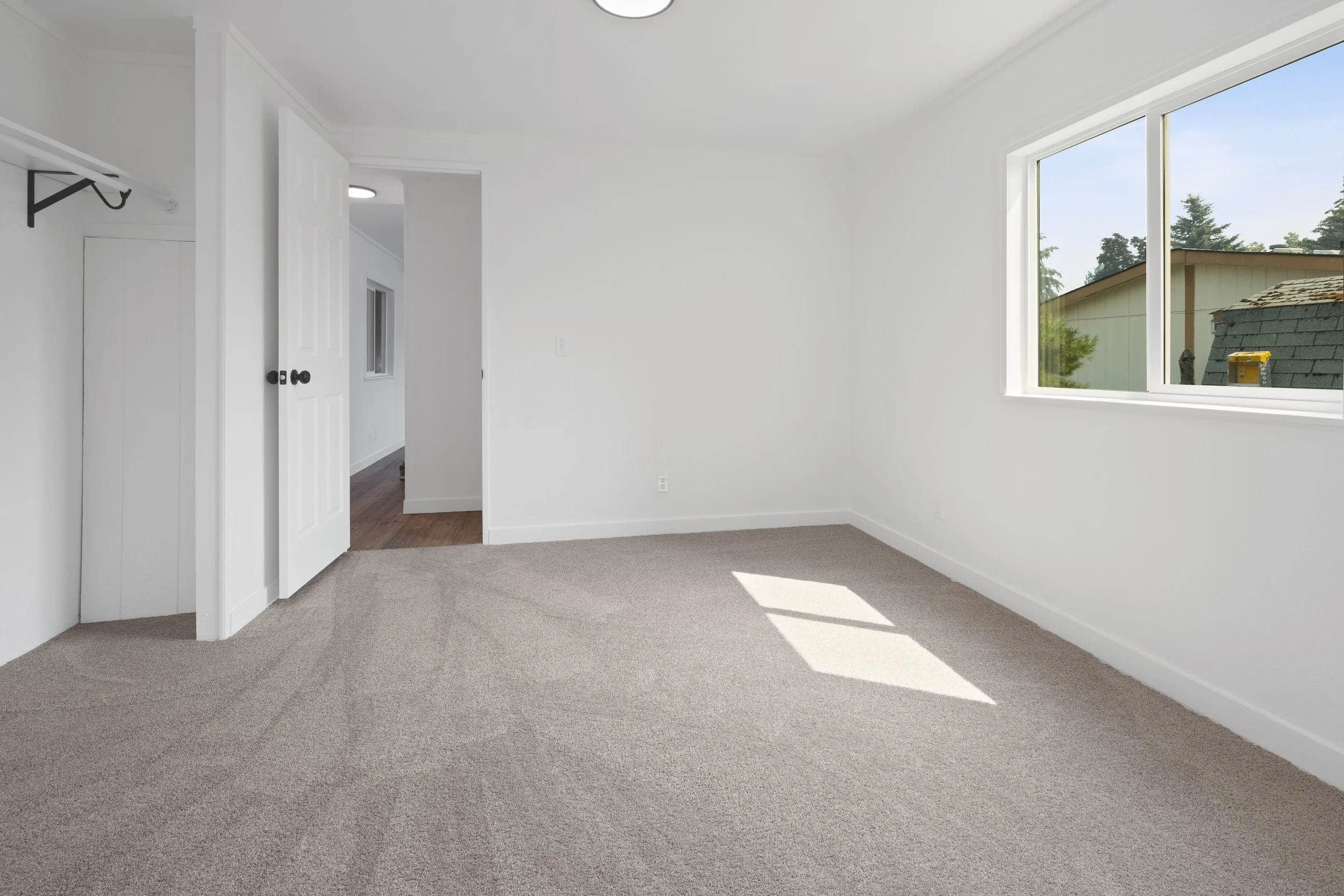 Empty room with beige carpet, white walls, a large window showing rooftops and trees, and an open door leading to a hallway.
