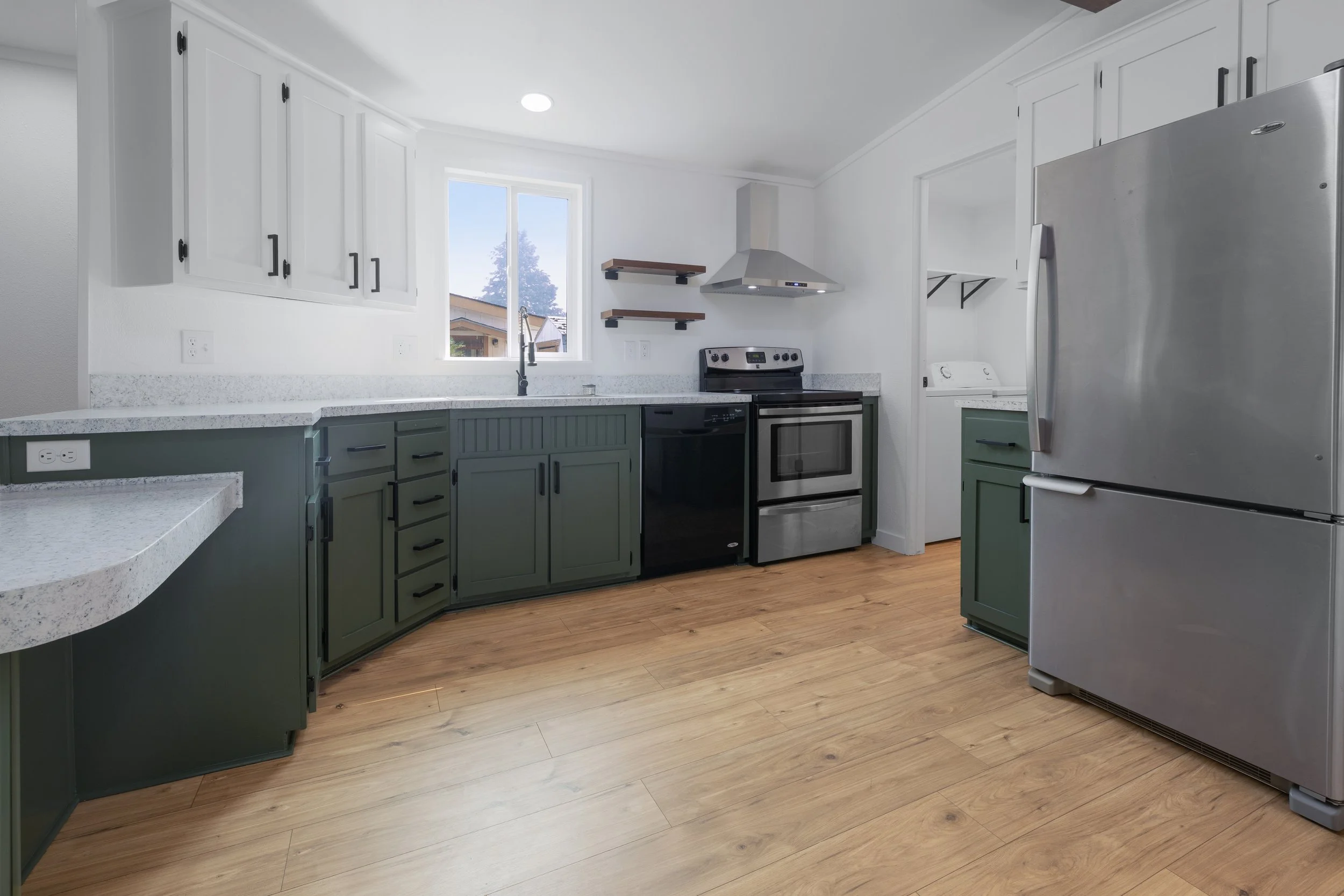 A modern kitchen with white upper cabinets, green lower cabinets, wood flooring, a stainless steel refrigerator, oven, and range hood. There is a window above the sink with a view of a neighboring house and trees.