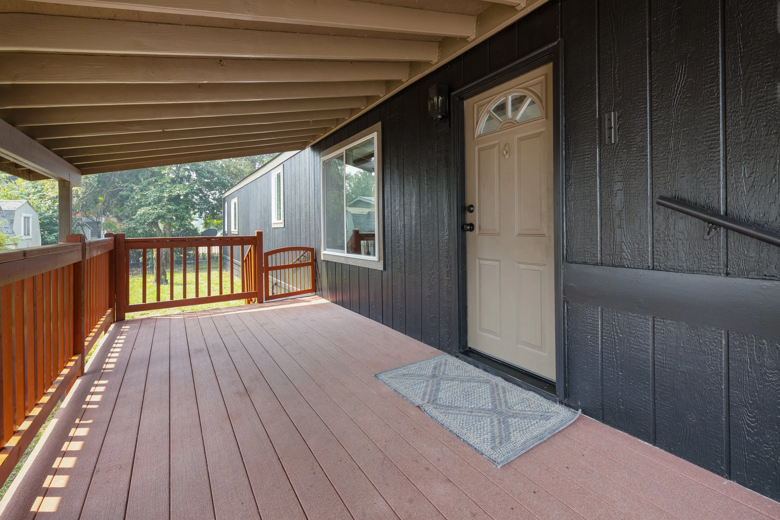 Porch with black exterior wall, beige door with a fan-shaped window, and wooden railing, with a view of trees and neighboring houses.