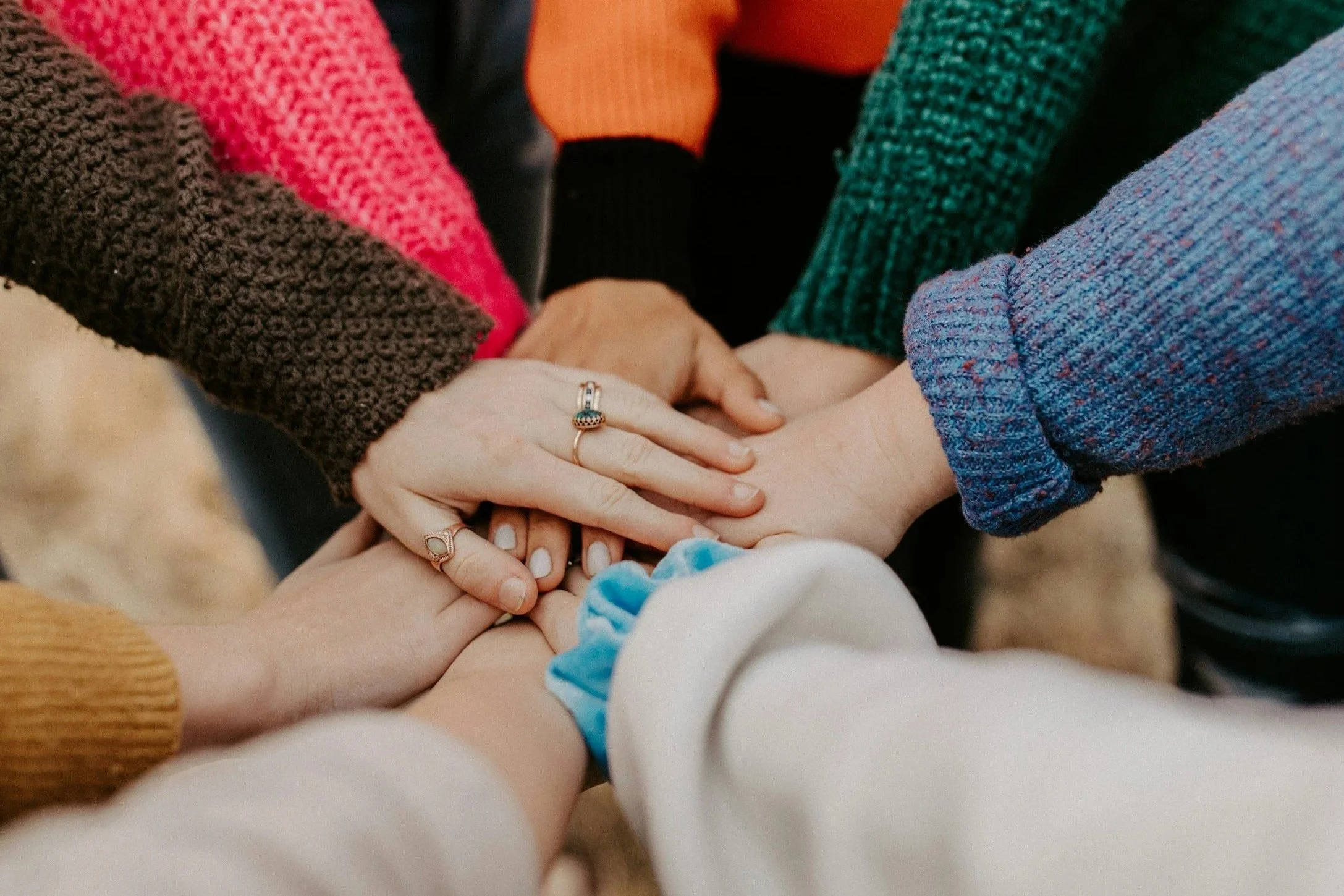 Close-up of multiple hands stacked together, showing diversity in skin tones and jewelry, with adults and a baby in colorful clothing, symbolizing unity and support.