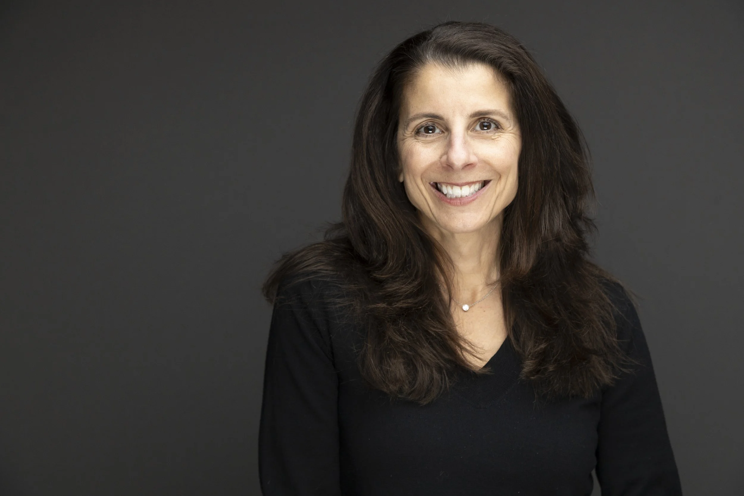 A woman with long brown hair, wearing a black top, smiling, against a dark gray background.