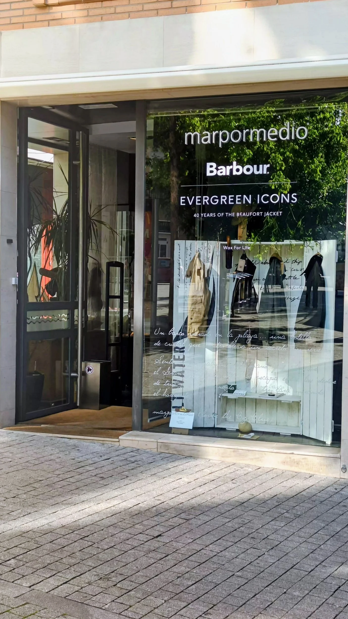Vista de la fachada de una tienda llamada 'marpormedio'. En la ventana se observan productos en exhibición y texto promocional, junto a un fondo que refleja árboles y la calle. La entrada es de vidrio con una puerta de doble hoja.