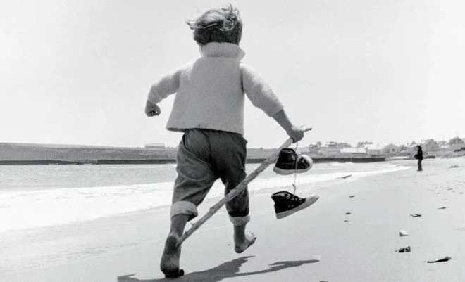 Niño caminando en la orilla del mar con sandalia en la mano y zapatos en el palo.