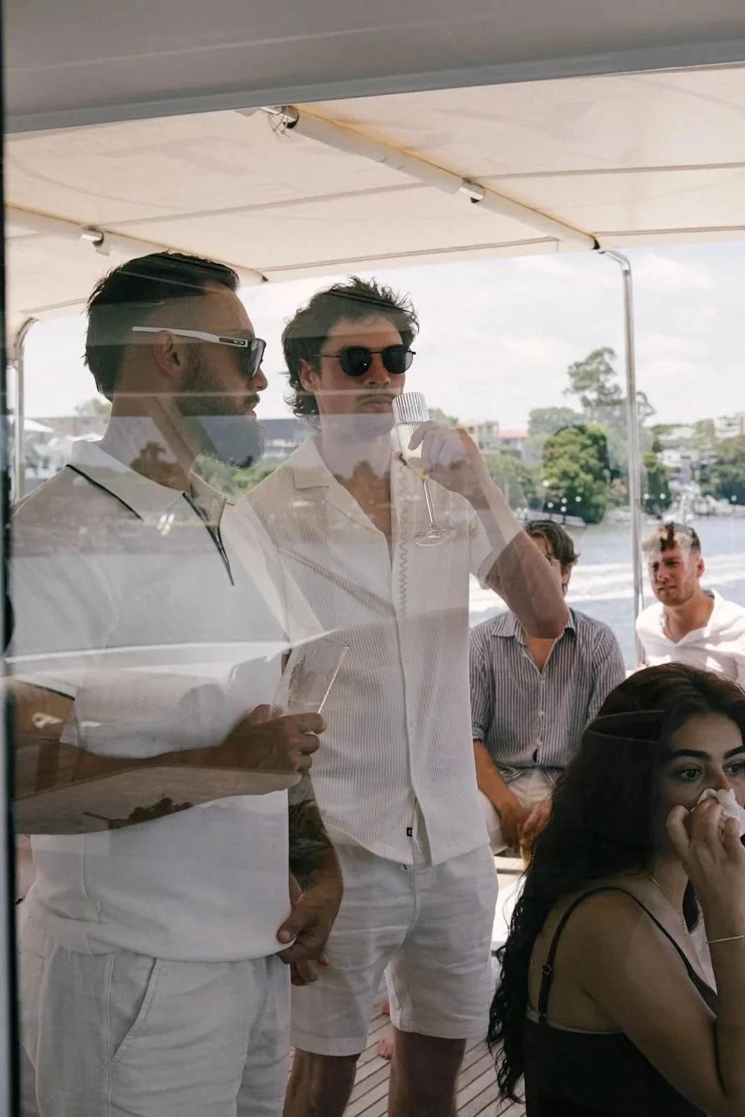Two men in white shirts and sunglasses are inside a boat, seen through a glass window. One is holding a microphone, and the other is holding a paper. Outside, people are seated on the deck, with water and trees in the background.