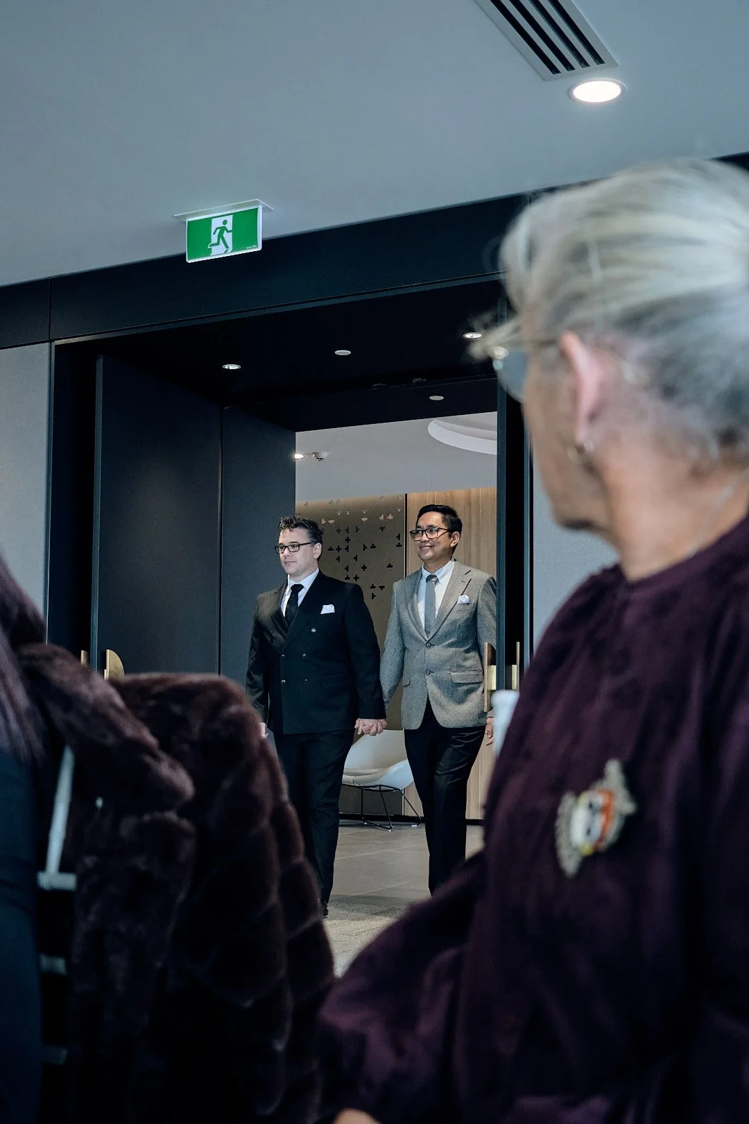 Two men in suits walking out of a room, holding hands, with audience members watching. Emergency exit sign above door.