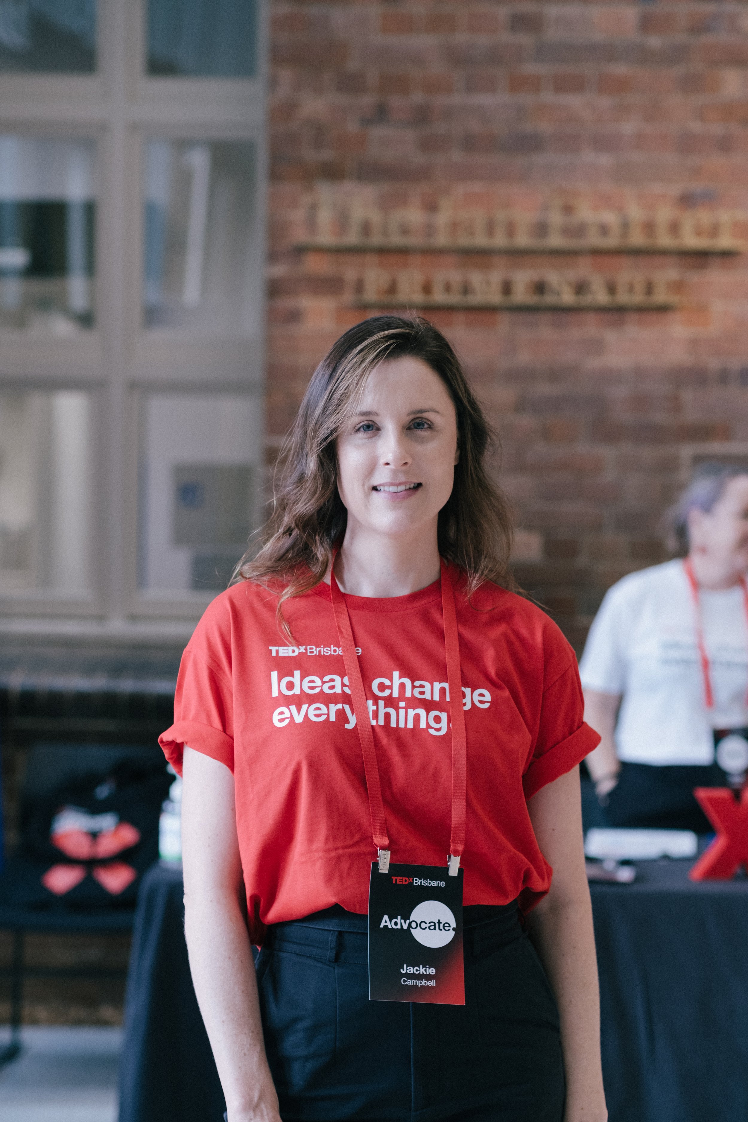A woman with shoulder-length brown hair and fair skin standing indoors, wearing a red TEDx Brisbane t-shirt with the slogan "Ideas change everything". She has a black lanyard with a visible name tag that reads "Jackie Campbell" and the WORD "Advocate