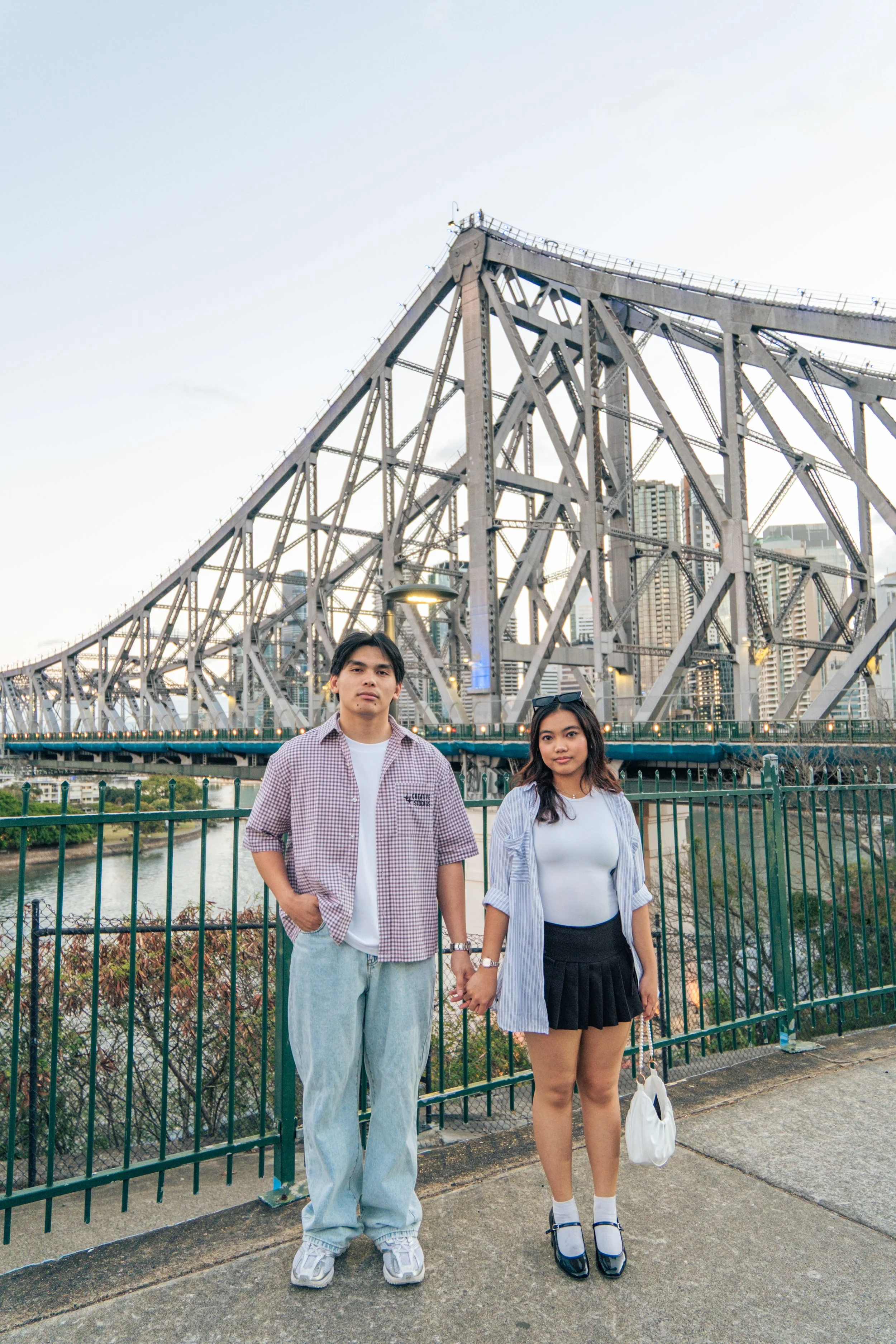 A man and a woman standing under a large bridge in an urban area, holding hands. The man is wearing a checkered shirt, white T-shirt, baggy jeans, and sneakers. The woman is dressed in a white top, striped long-sleeve shirt, black skirt, and Mary Jane shoes, holding a white bag. Buildings are visible in the background.