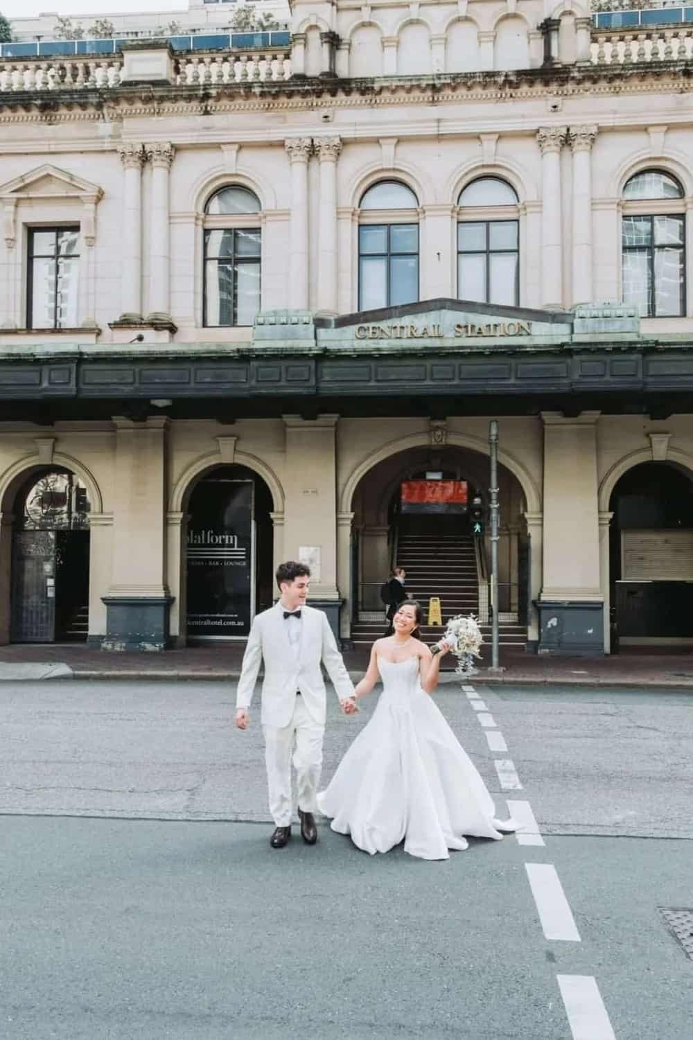 A newlywed couple in wedding attire walking hand in hand in front of a historic building labeled 'Central Station'. The bride is holding a bouquet and smiling, while the groom looks at her. The scene appears joyful and festive.