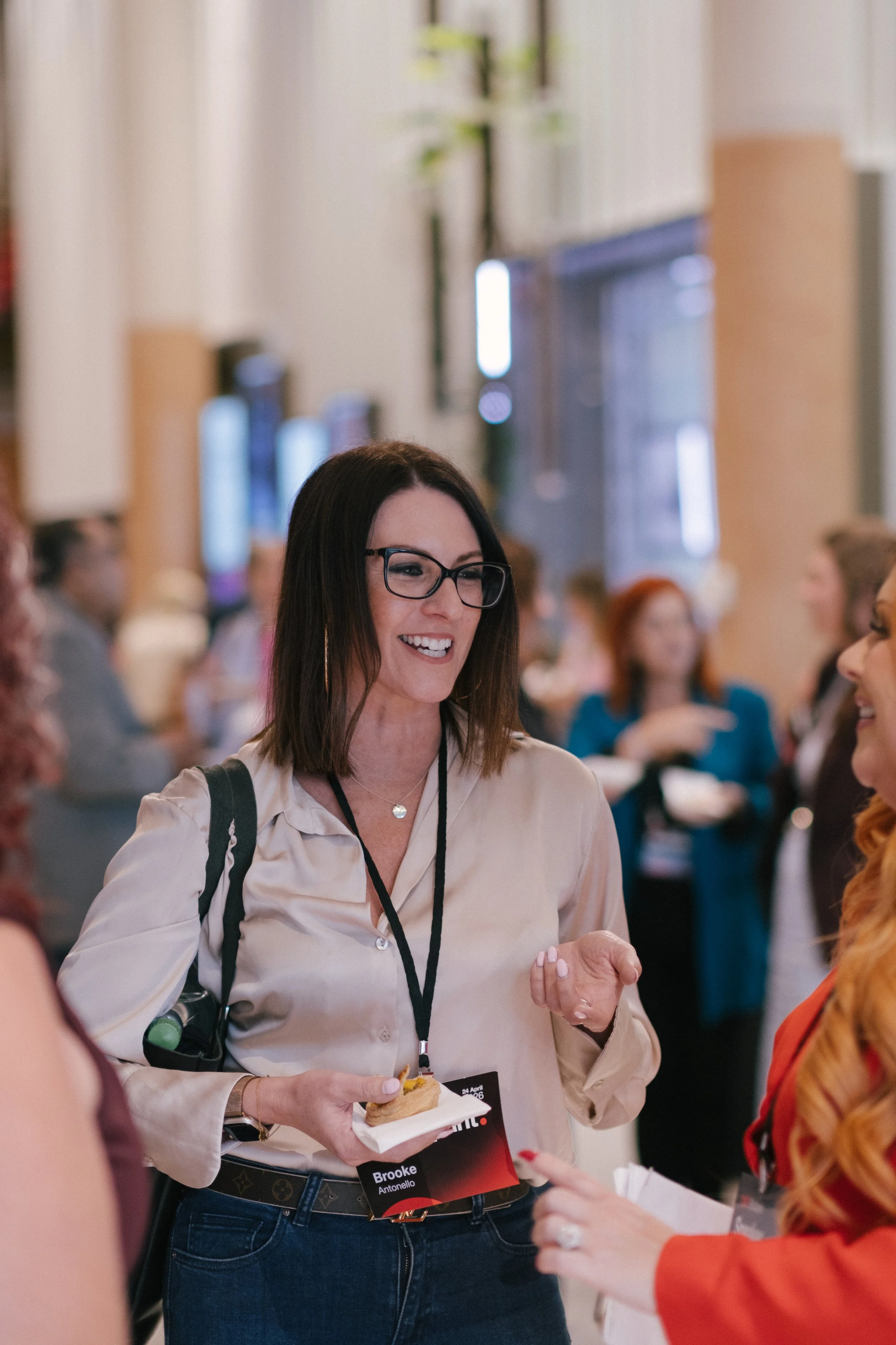 A woman with dark brown hair, glasses, and a white shirt smiling while talking to another person at a crowded indoor event. She is holding a small plate with food and has a name tag that reads "Brooke Antonello."