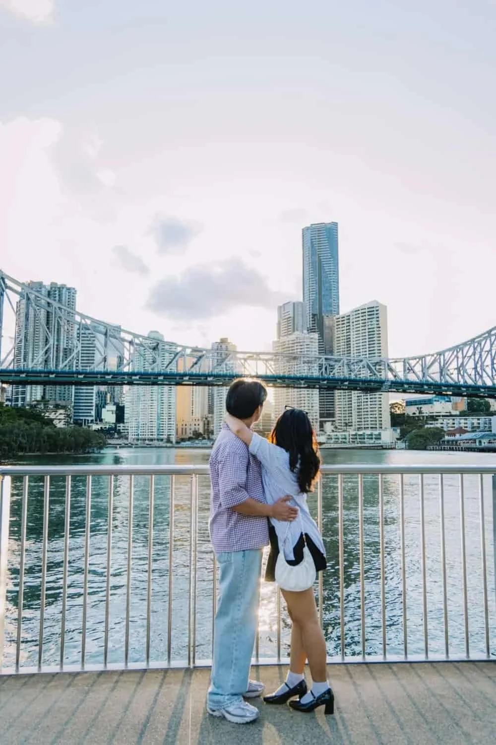 A couple standing by a railing on a riverbank at sunset, with a city skyline and bridge in the background.