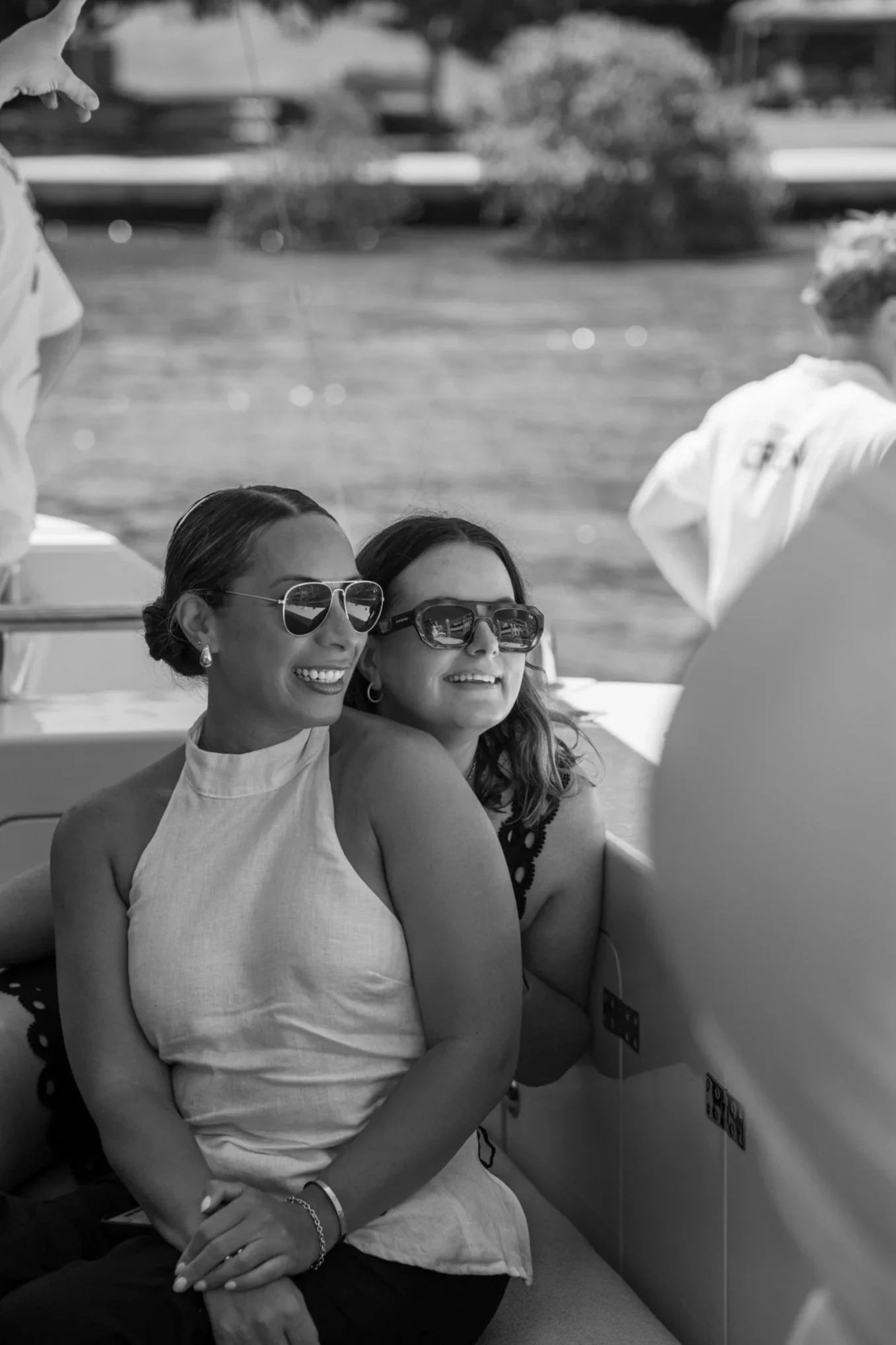 Two women smiling and wearing sunglasses, sitting on a boat with a water background, with a man in a hat standing nearby.