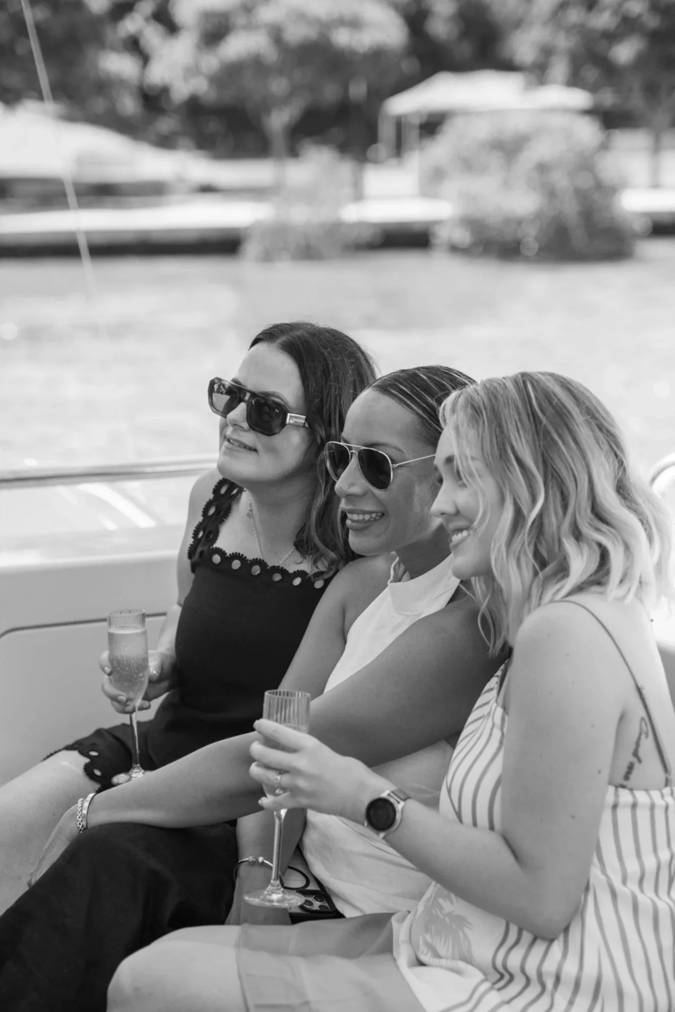 Three women sitting side by side on a boat, holding glasses of champagne, smiling, wearing sunglasses, with a blurred background of trees and a gazebo.