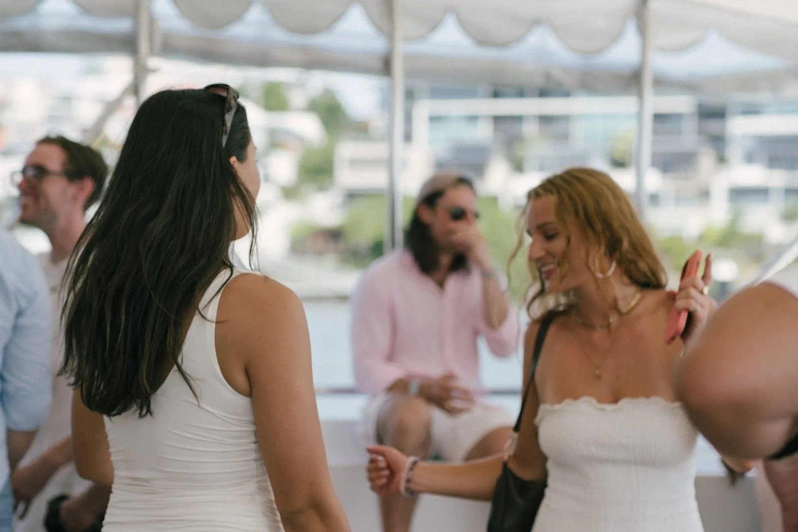 A group of people socializing at an outdoor event under a tent, including two women in white tops, one with long dark hair and sunglasses on her head, and the other with curly red hair holding a pink item, smiling and talking with the others in the b