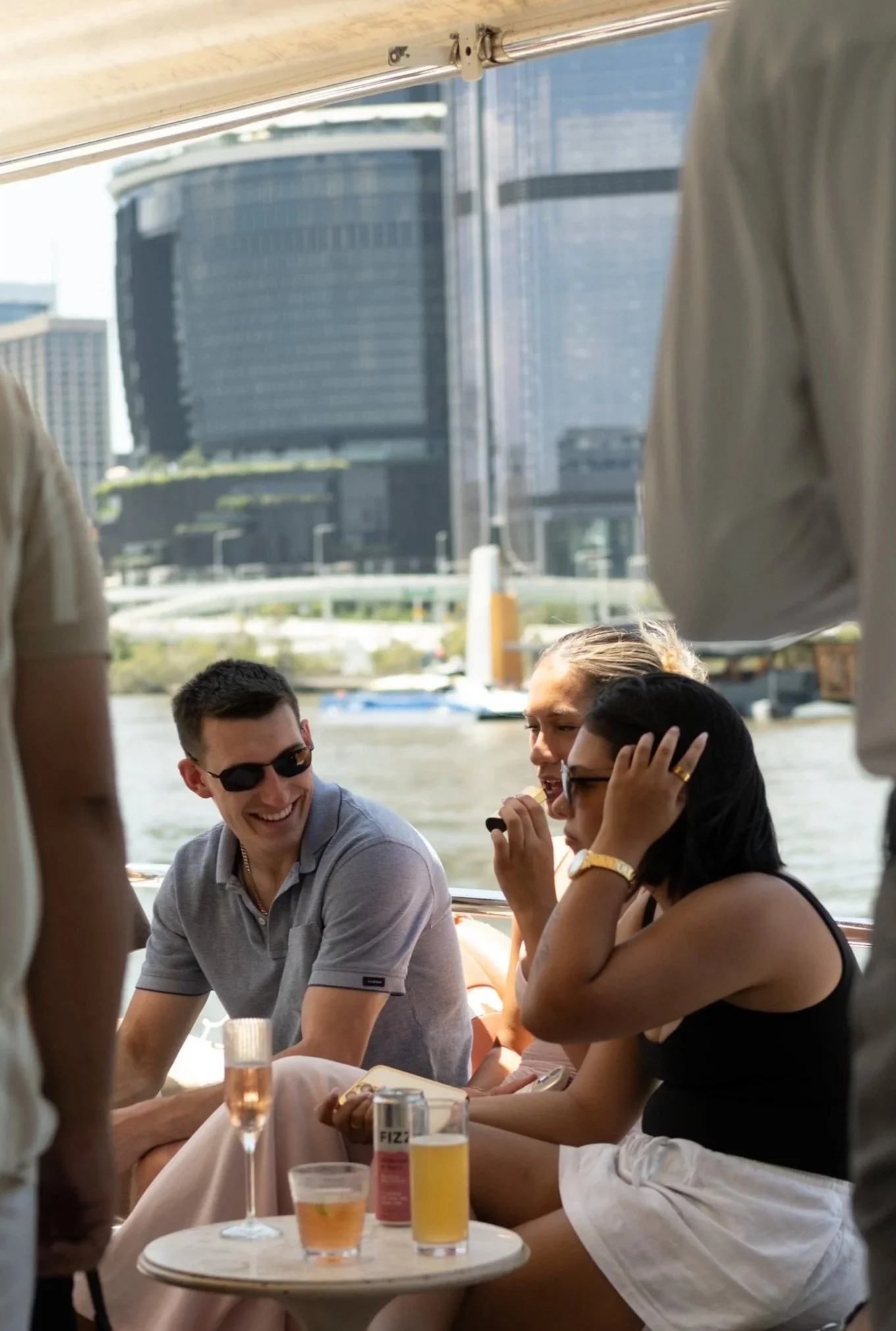 A group of people sitting outdoors by the water, enjoying drinks and conversations with a city skyline in the background.