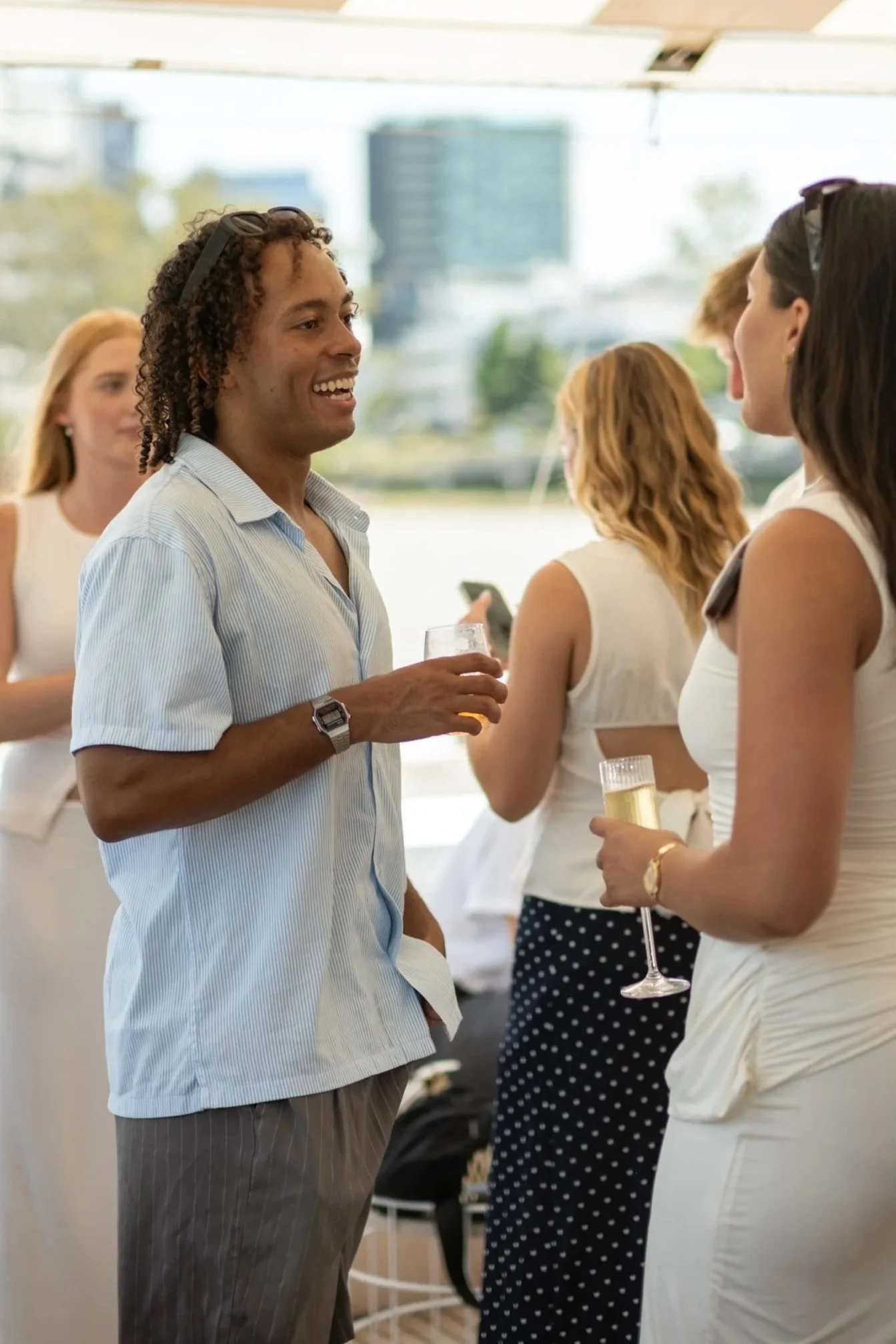 People at a social gathering outdoors, engaging in conversation, with two women holding glasses of champagne and a man smiling while talking to one woman.