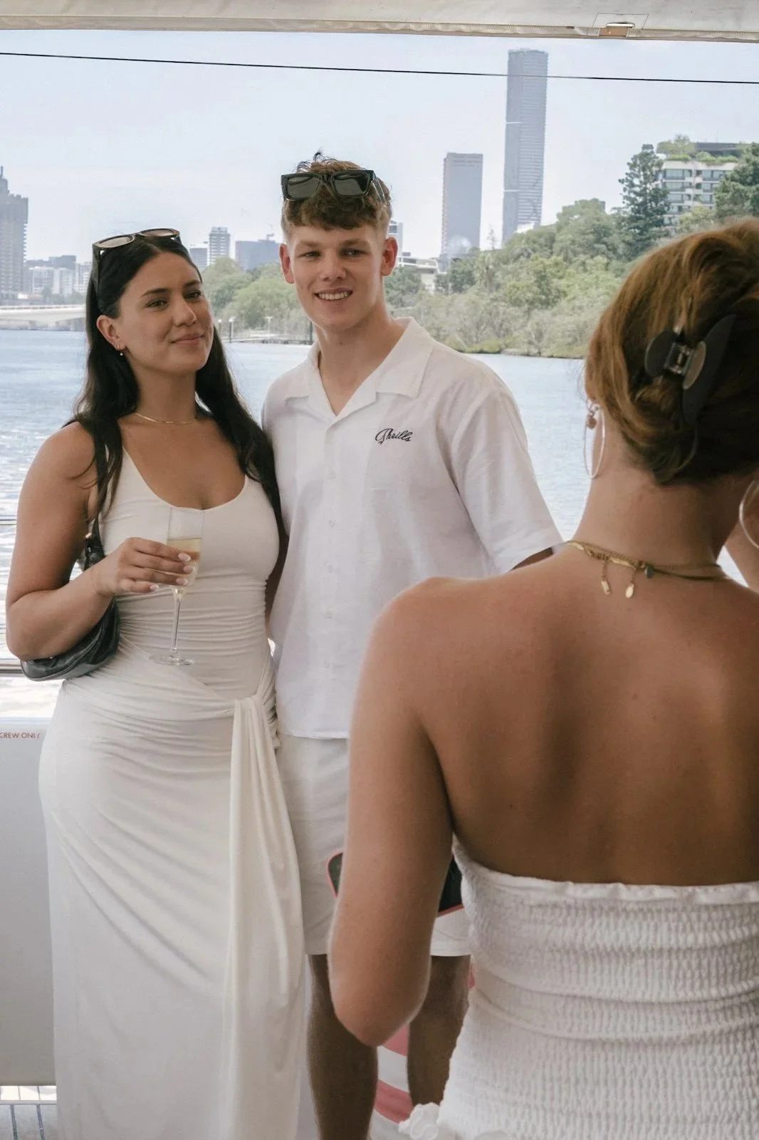 People at a social event near a body of water with a city skyline in the background, including tall buildings and trees, during daytime.