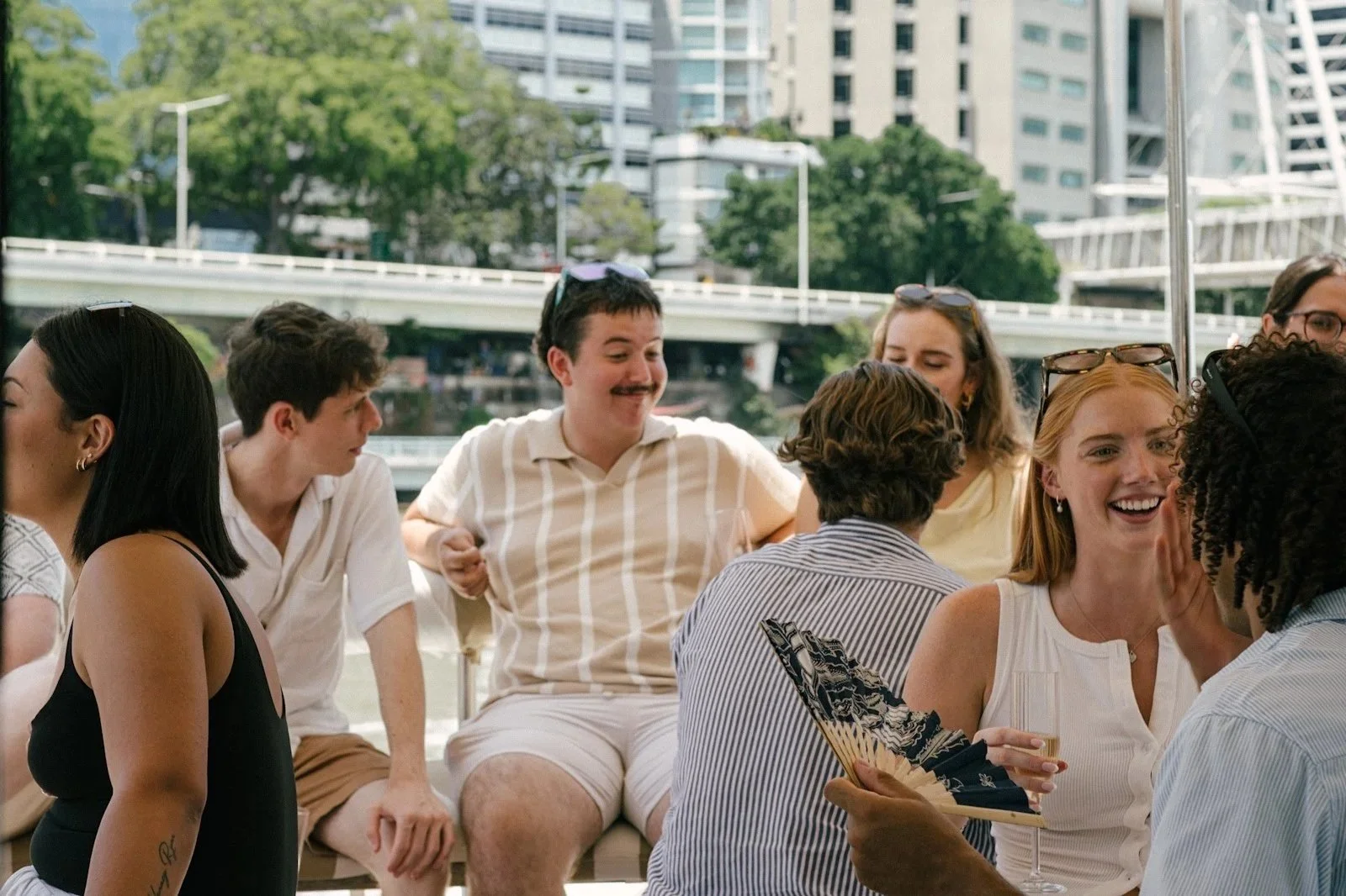 A group of people sitting outdoors on a boat, smiling and chatting. The background shows an urban cityscape with trees, buildings, and a bridge.