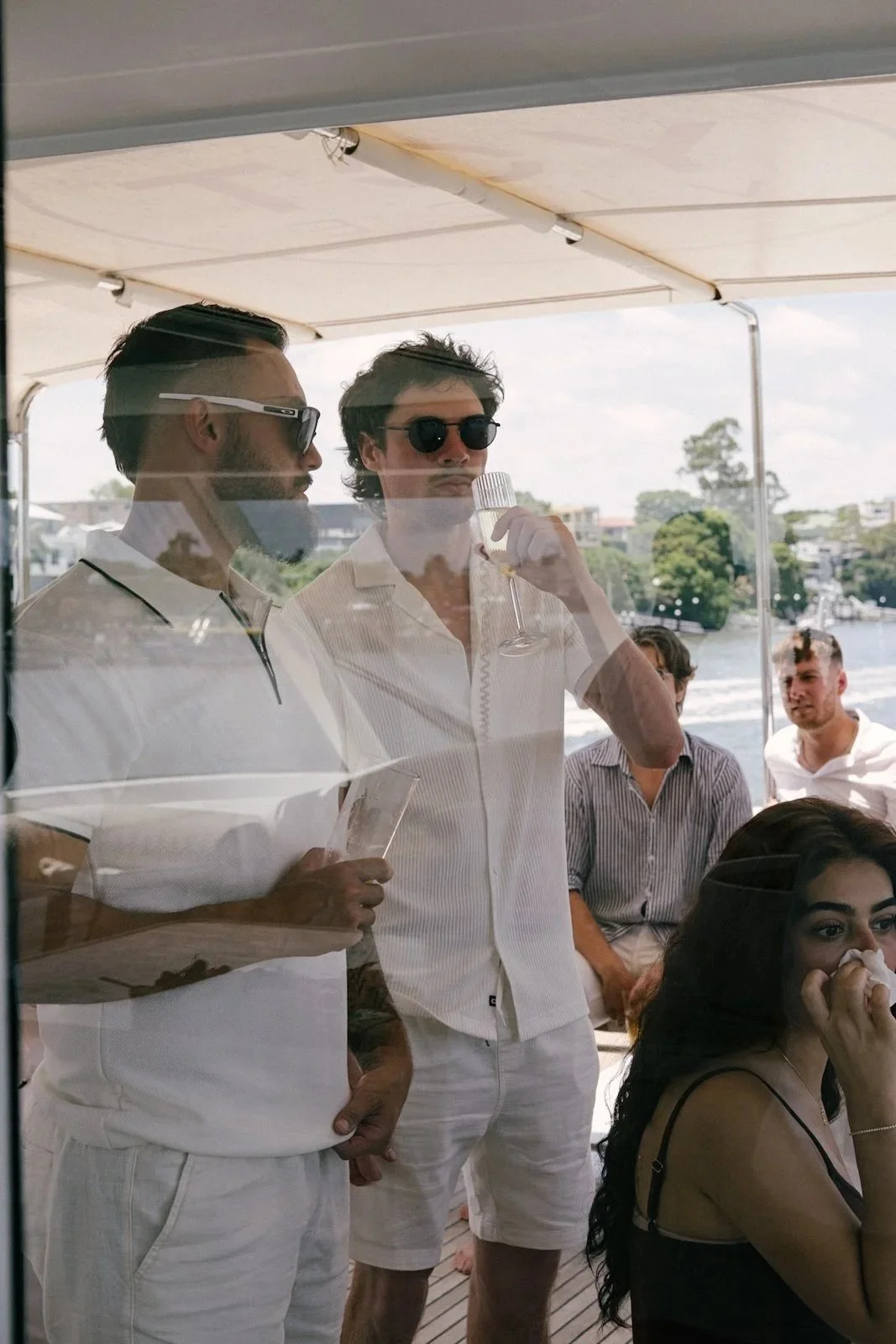 People on a boat with a view of a river, trees, and buildings in the background, some wearing sunglasses, one drinking from a glass, during the daytime.