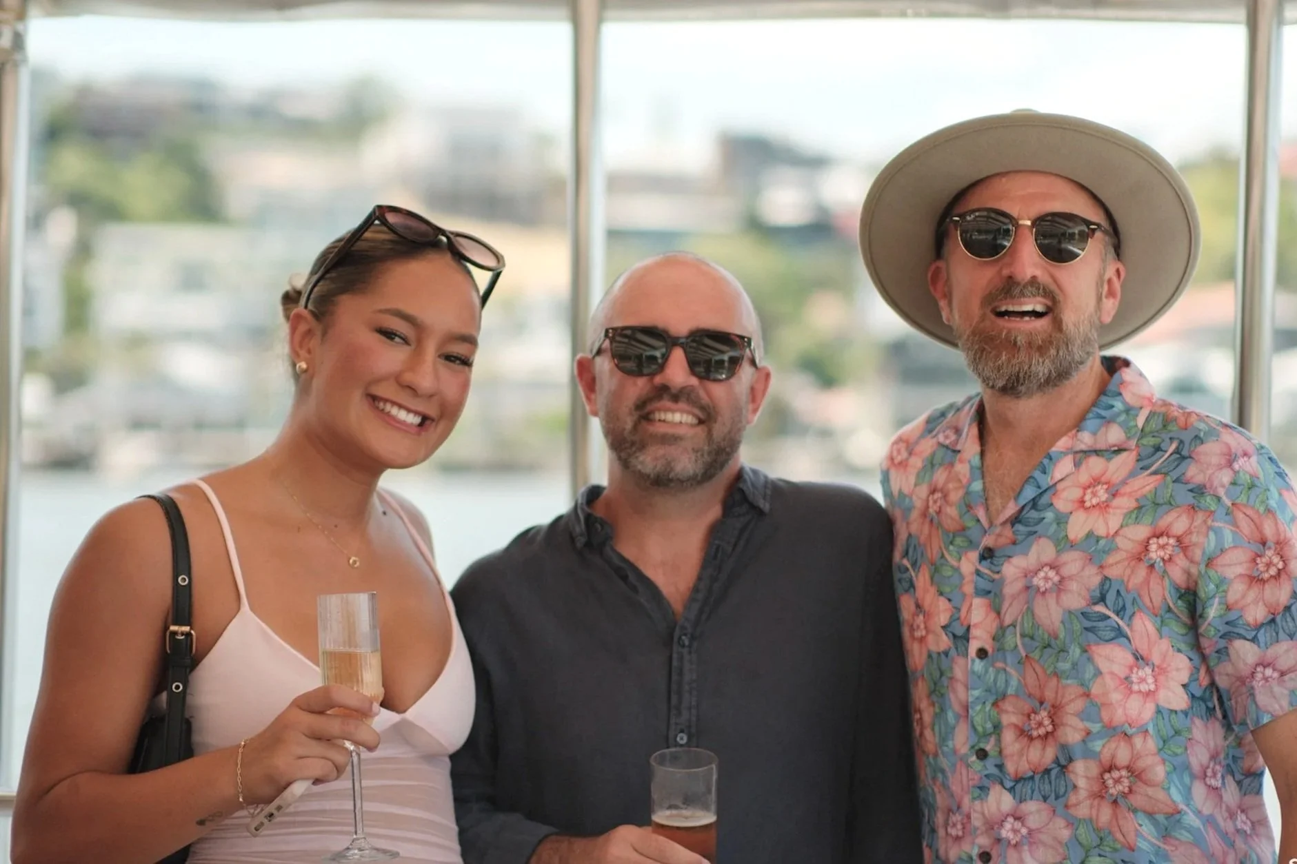 Three adults at a social gathering, dressed in summer attire, holding drinks, smiling, standing indoors with a view of a river or canal in the background.