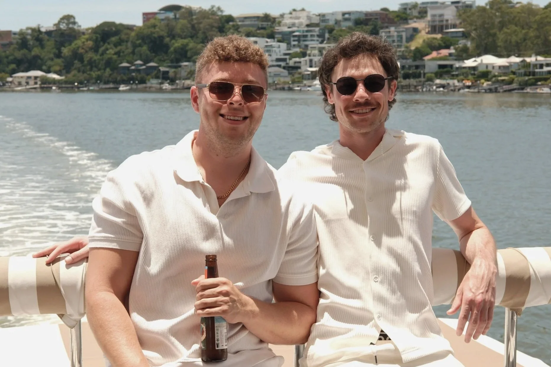 Two men smiling and wearing sunglasses sitting on a boat with a waterway and hillside houses in the background.