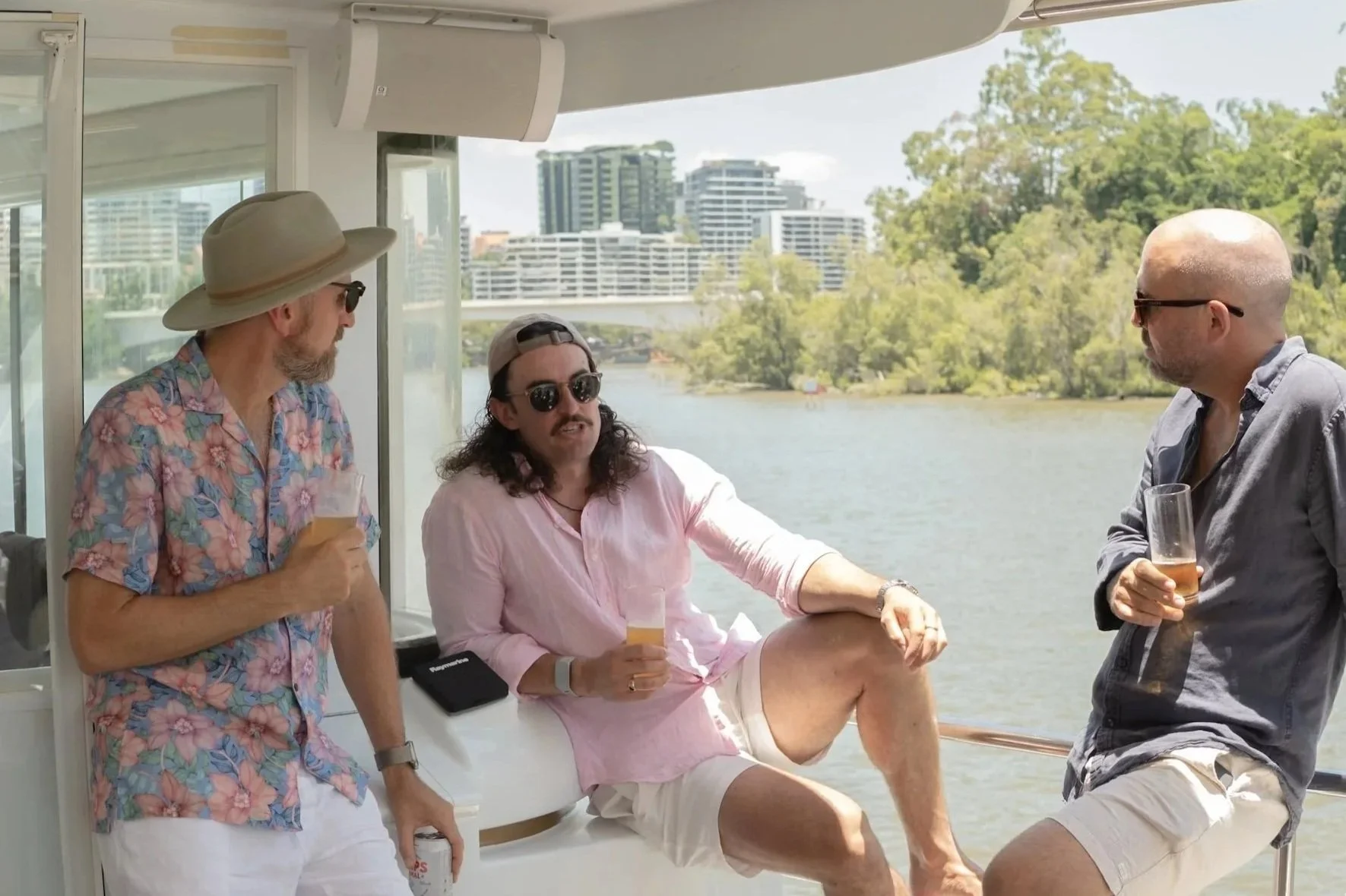 Three men on a boat at a river, casually dressed, holding drinks, with city buildings and greenery in the background.