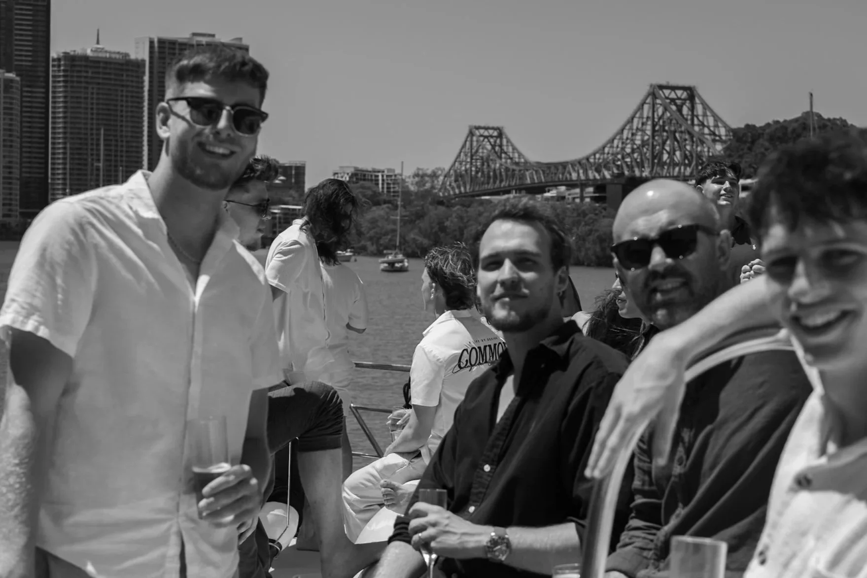 Group of people enjoying drinks outdoors by a river with a city skyline and a bridge in the background.