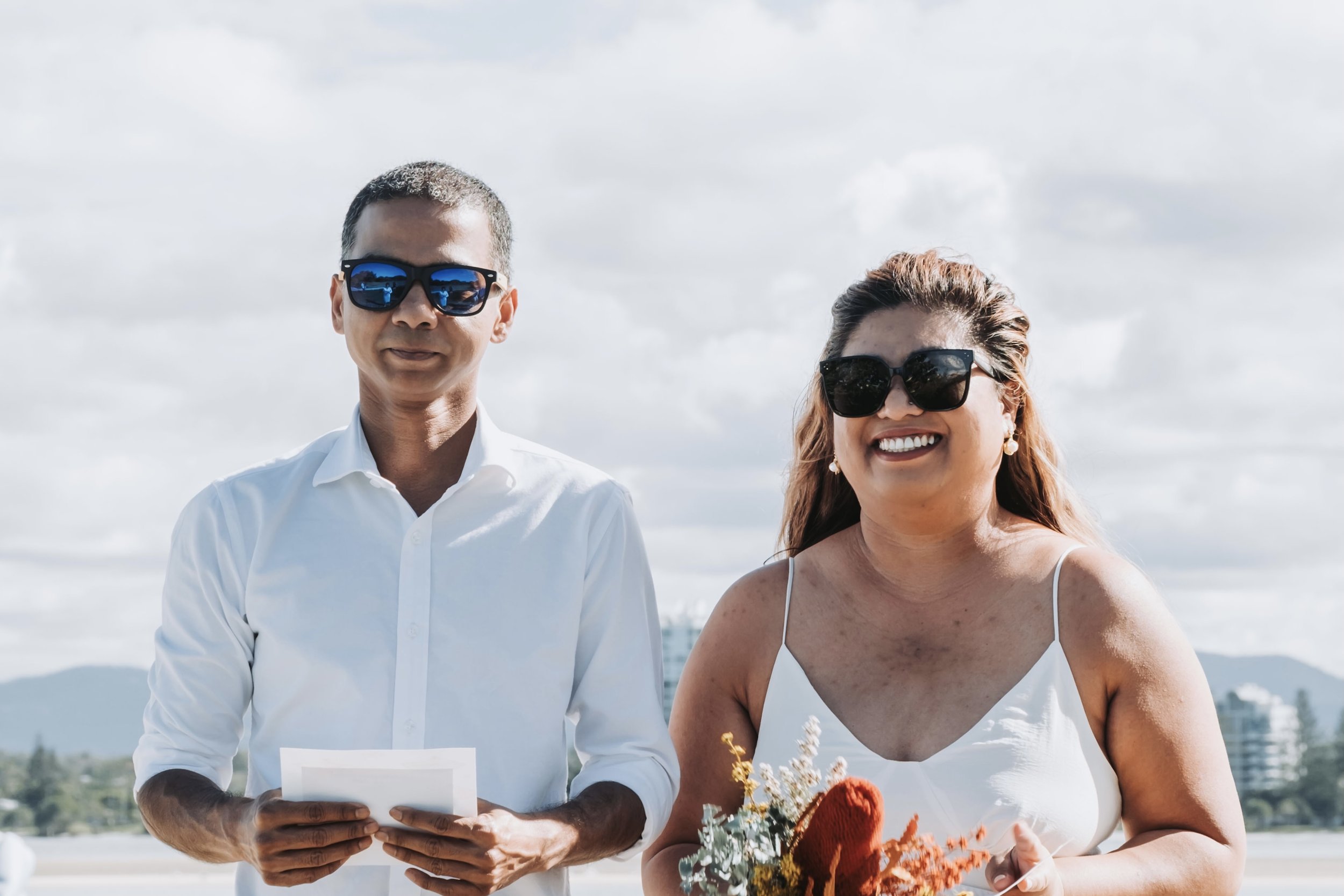 A man and a woman standing outdoors near the water on a cloudy day, both wearing sunglasses. The woman is smiling and holding a bouquet of flowers, and the man is holding a piece of paper.