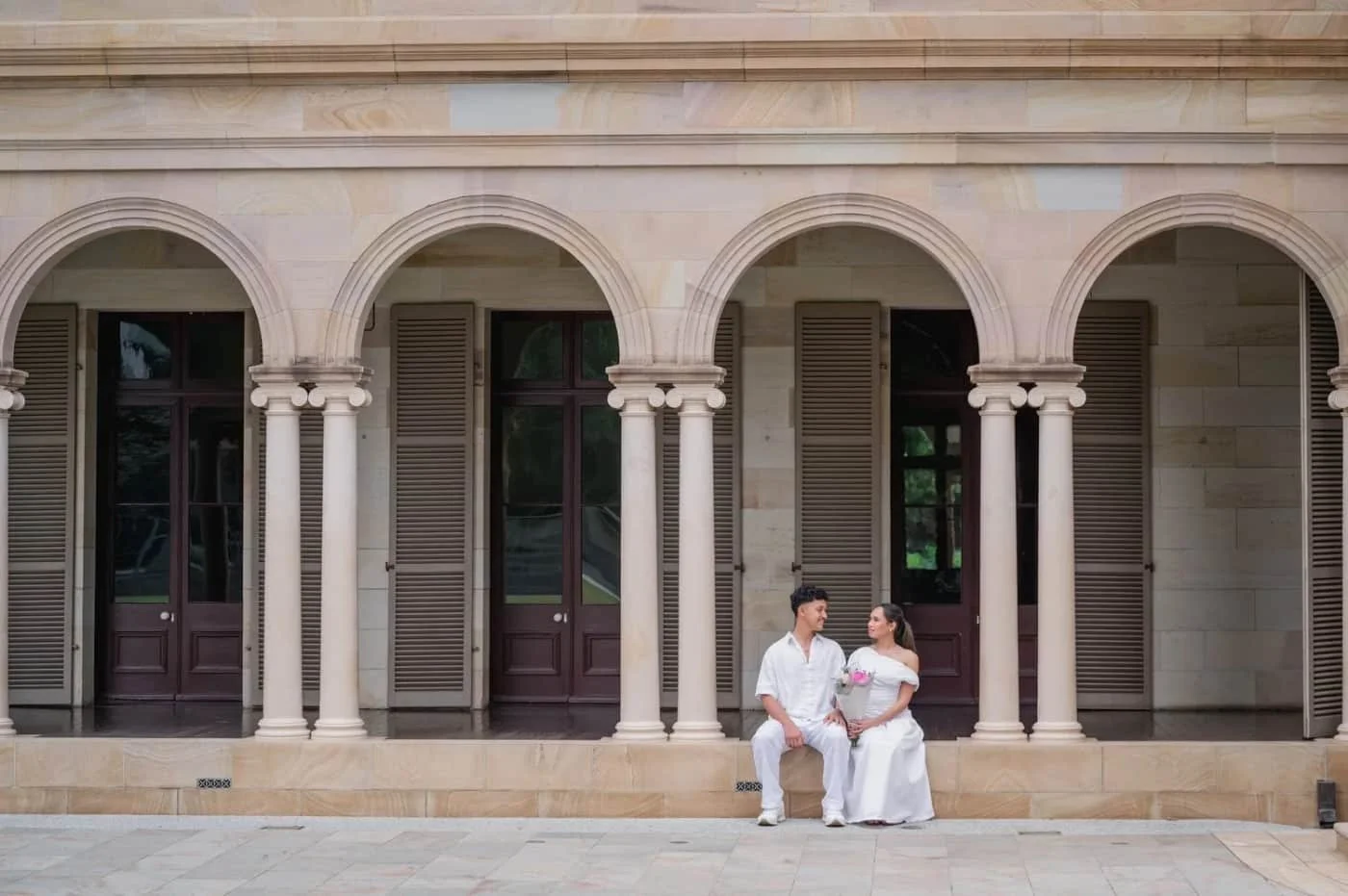 A couple dressed in white sitting on a step in front of a building with arches and wooden shutters, engaged in conversation.