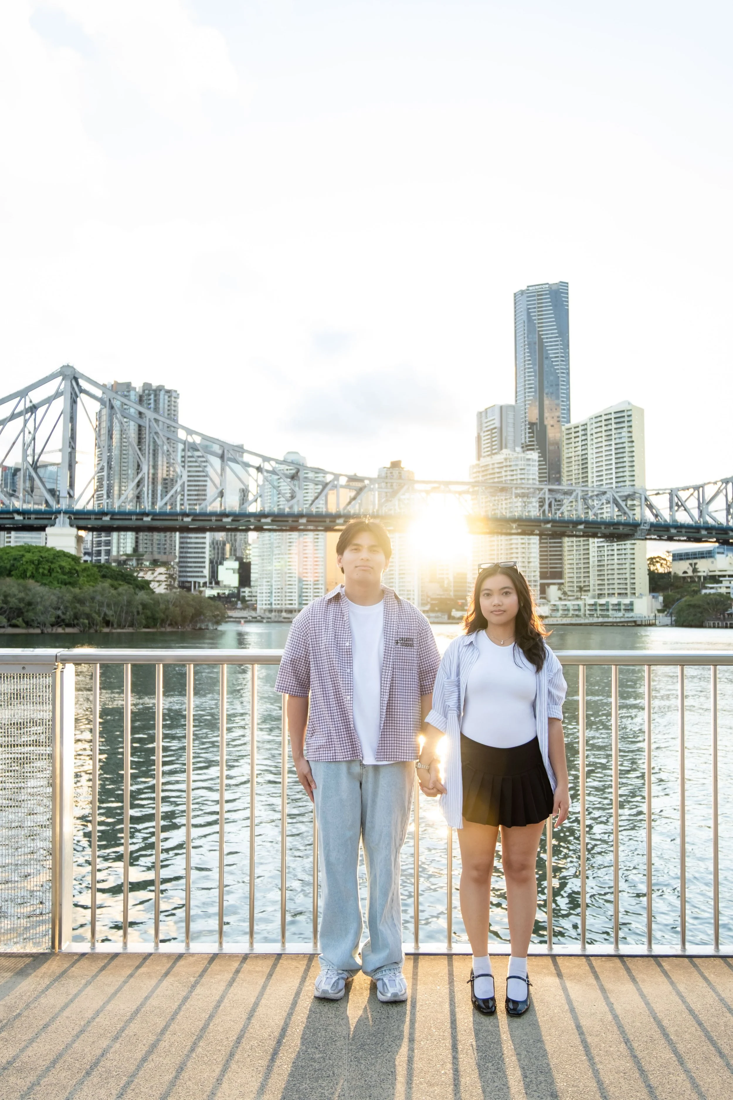 A young man and woman standing hand-in-hand on a bridge by a river in a city with tall modern skyscrapers, during sunset.