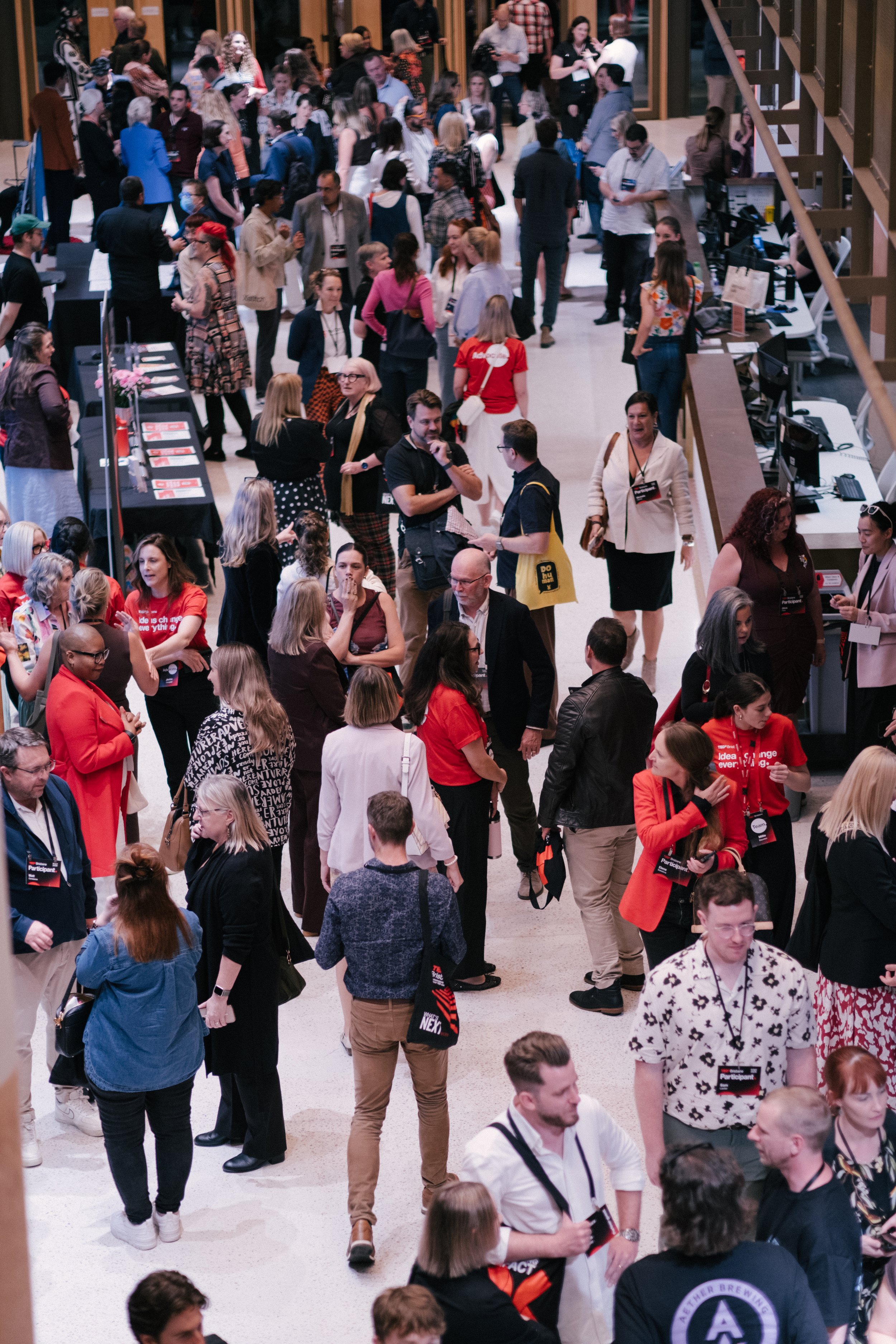 A busy conference or networking event with numerous people walking and talking, some wearing conference badges and lanyards, in a large hall with booths and displays.