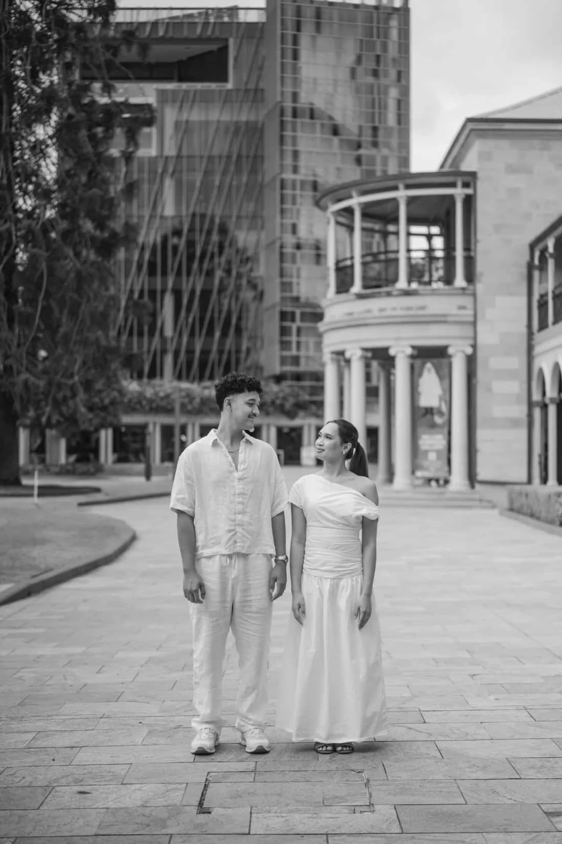 A black and white photo of a young man and woman walking and smiling at each other on a city street with modern buildings in the background.