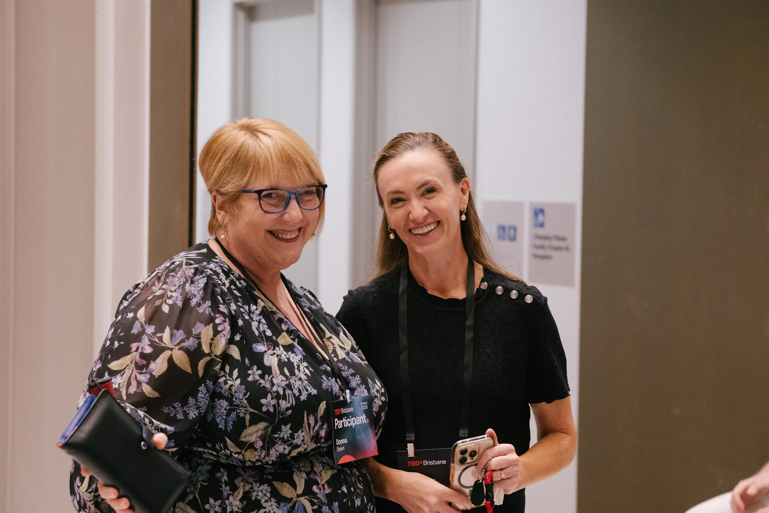 Two women smiling, standing close together at a conference or event, holding phones and wearing badges with 'TEDx Brisbane' on them.