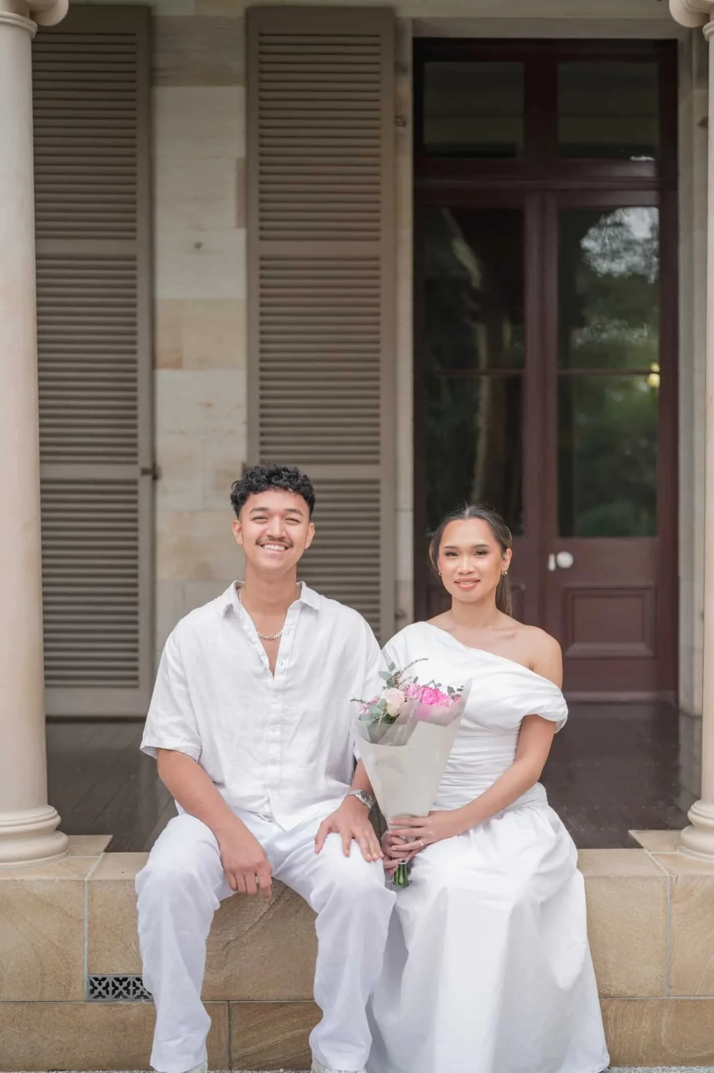 Couple in white sitting on outdoor stairs with a pink bouquet