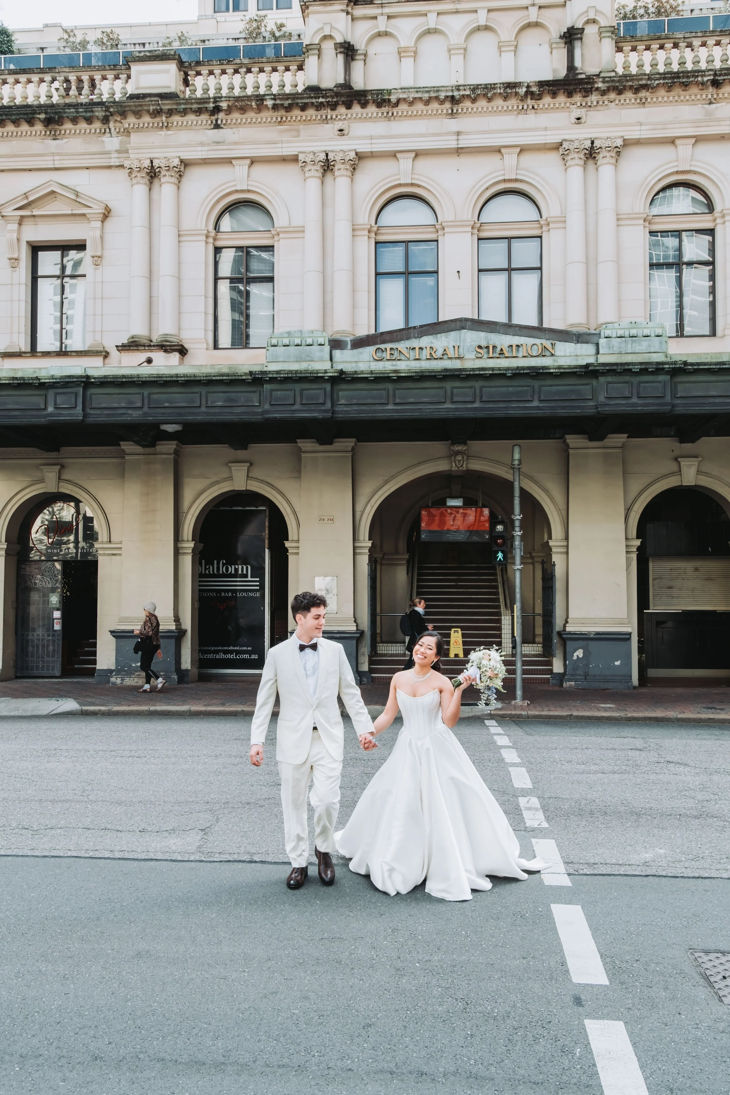 Bride and groom walk hand-in-hand across a street in front of Central Station in elegant wedding attire.
