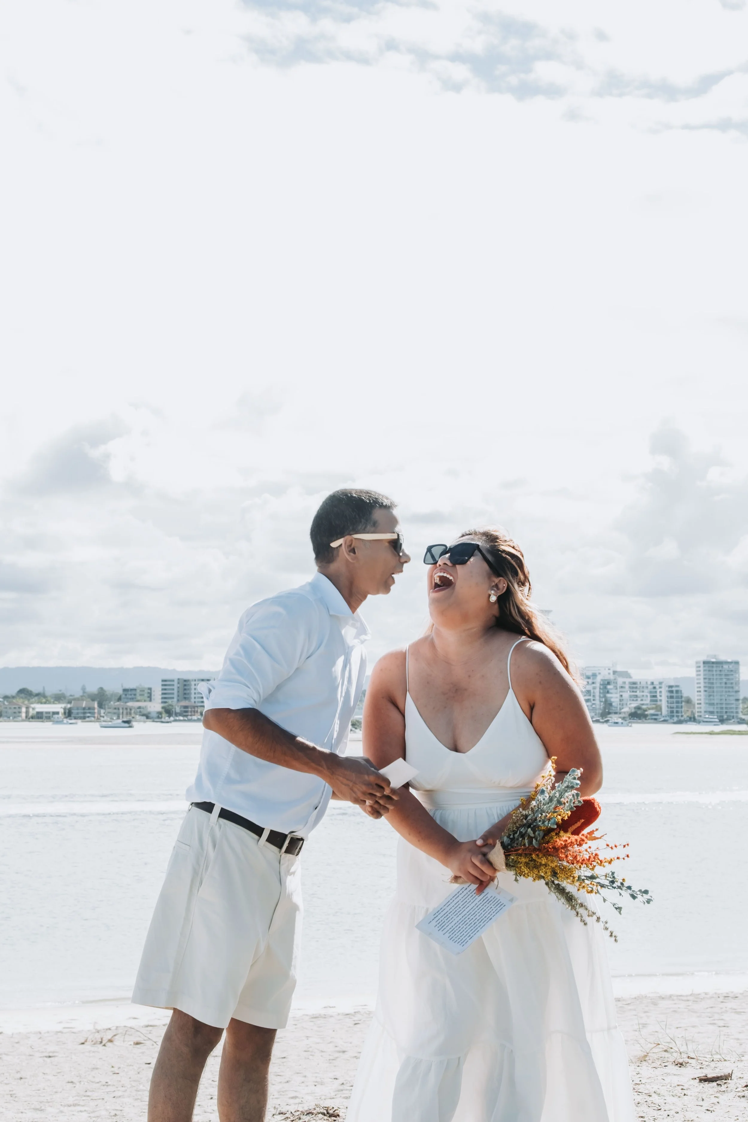 A couple in wedding attire on a beach, laughing and holding hands, with a city skyline in the background.