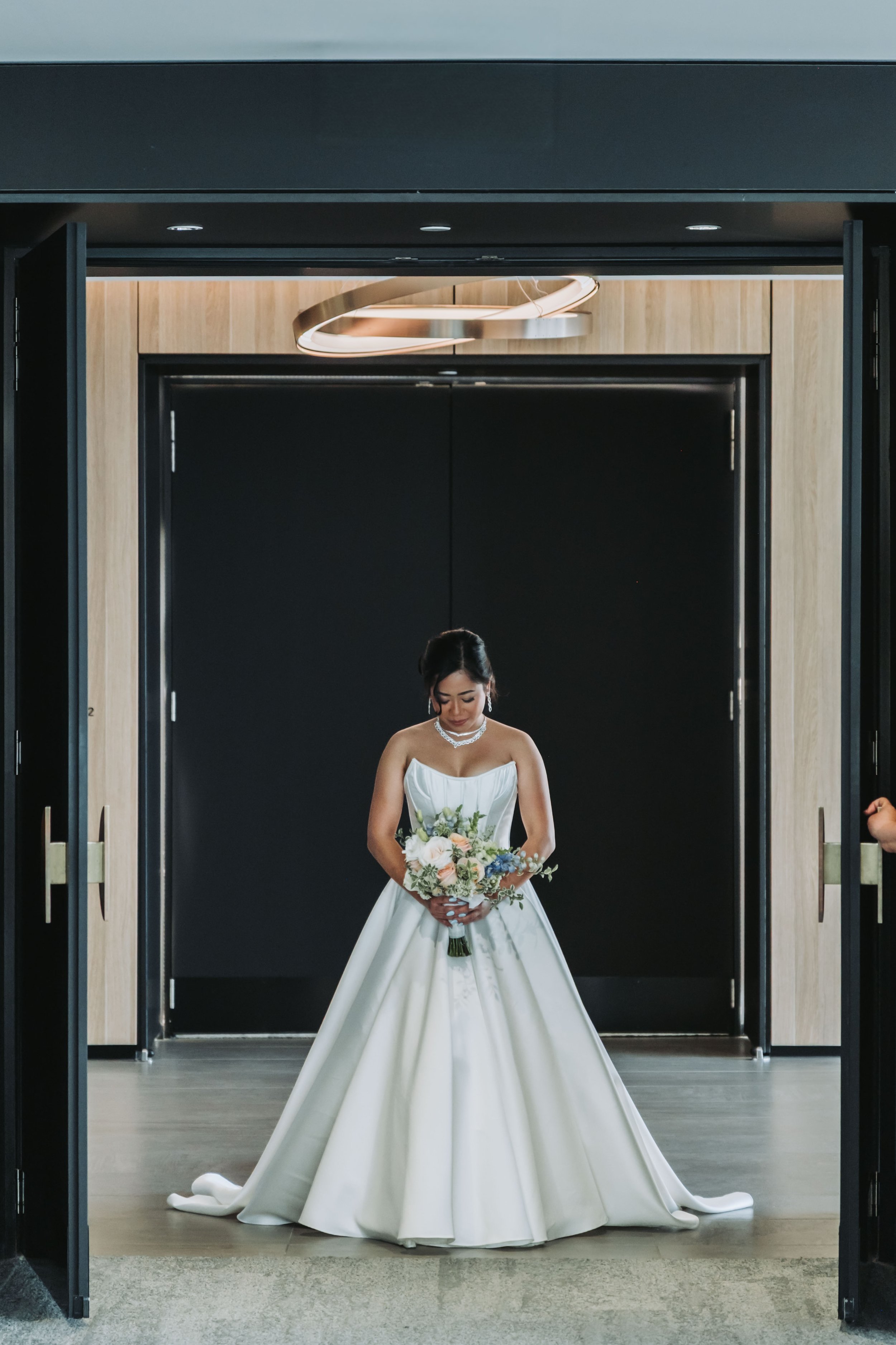 A bride in a strapless white wedding gown holding a bouquet of flowers, standing in a doorway with black and wooden walls.