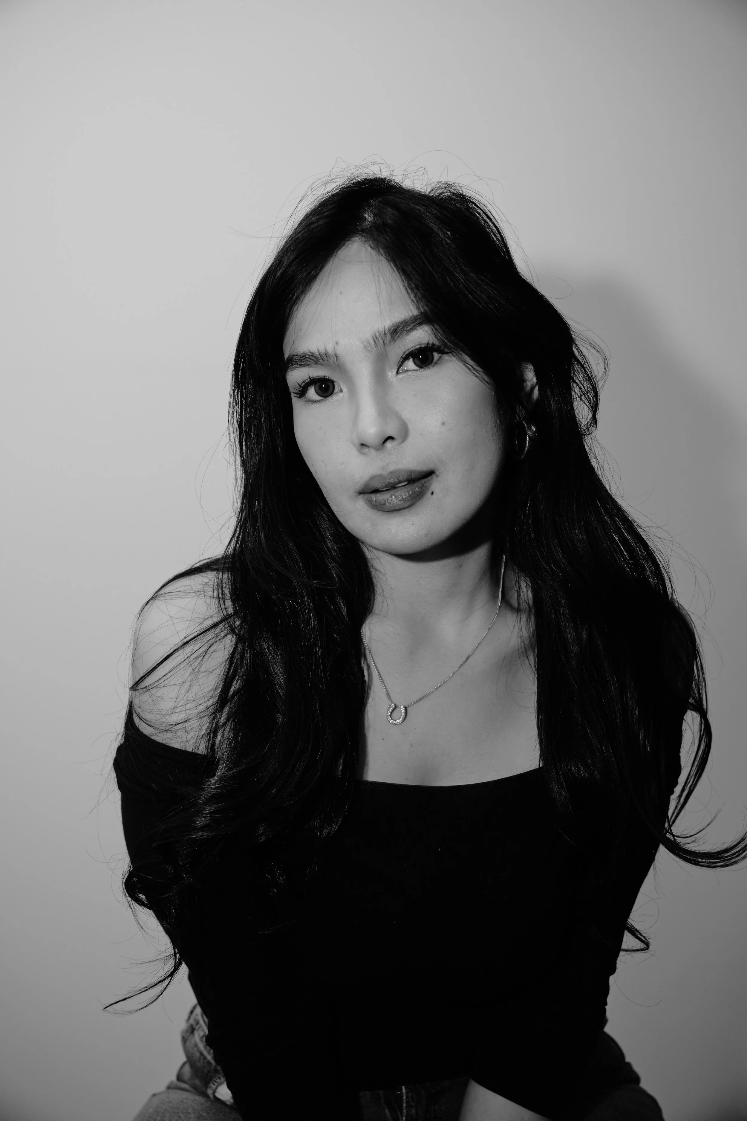Black and white portrait of a woman with long wavy hair, looking directly at the camera, wearing a black off-the-shoulder top and a necklace with a circular pendant.
